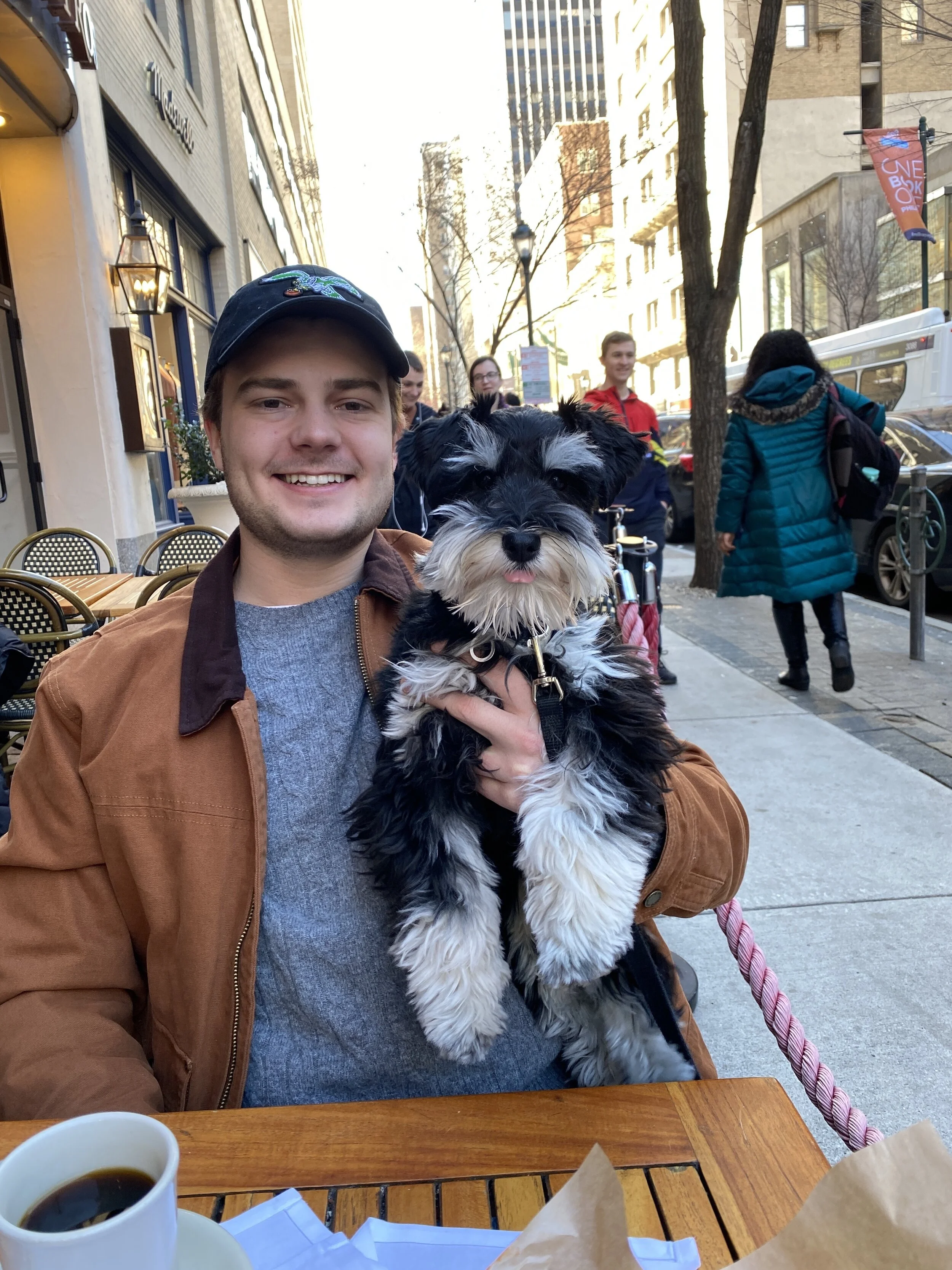 A man wearing a baseball cap smiling and holding a black and white dog with its tongue sticking out, seated at an outdoor cafe table with coffee, in an urban street scene with people walking by and tall buildings in the background.