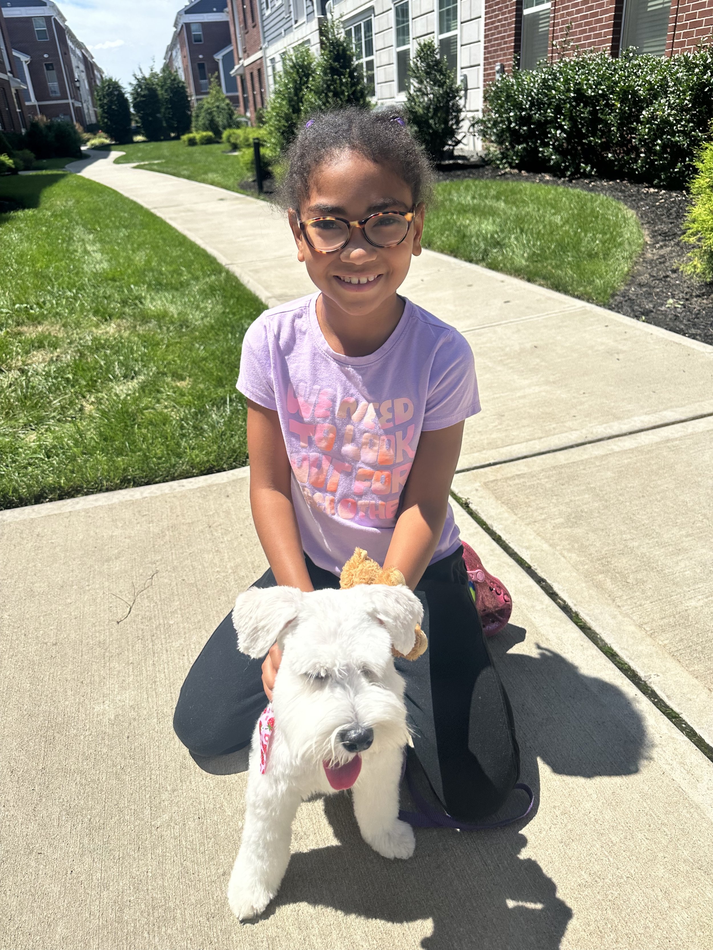 A smiling young girl with glasses and a pink T-shirt crouching on a sidewalk, holding a stuffed dog toy. A large fluffy white dog with black eyes and nose is in front of her, facing the camera. The background shows a residential complex with colorful