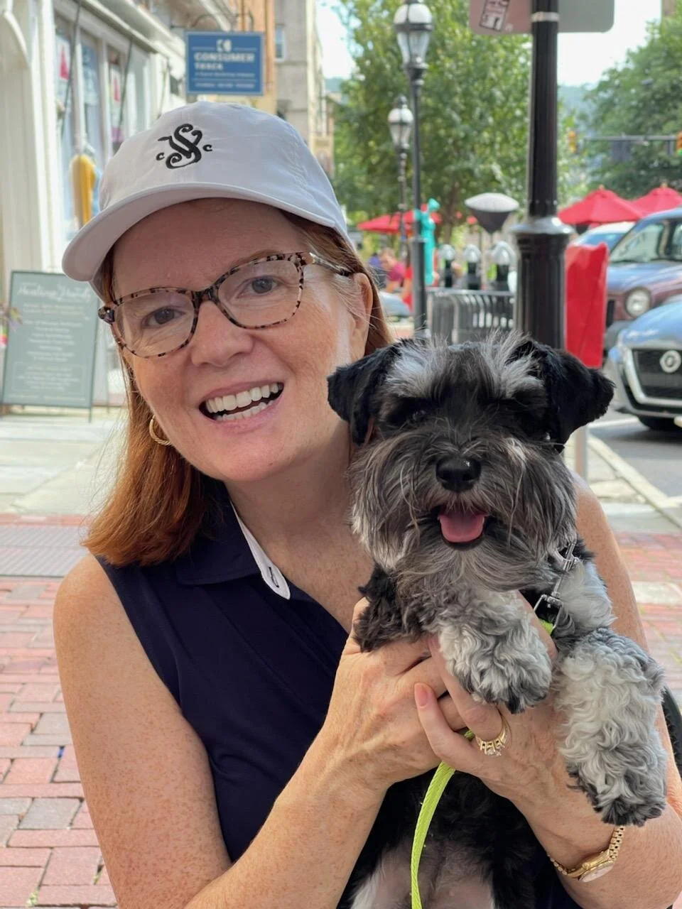 A woman with red hair, wearing glasses, a white cap, and a navy sleeveless top, smiling while holding a small black and gray puppy with a fluffy coat and a happy expression, outside on a city sidewalk.