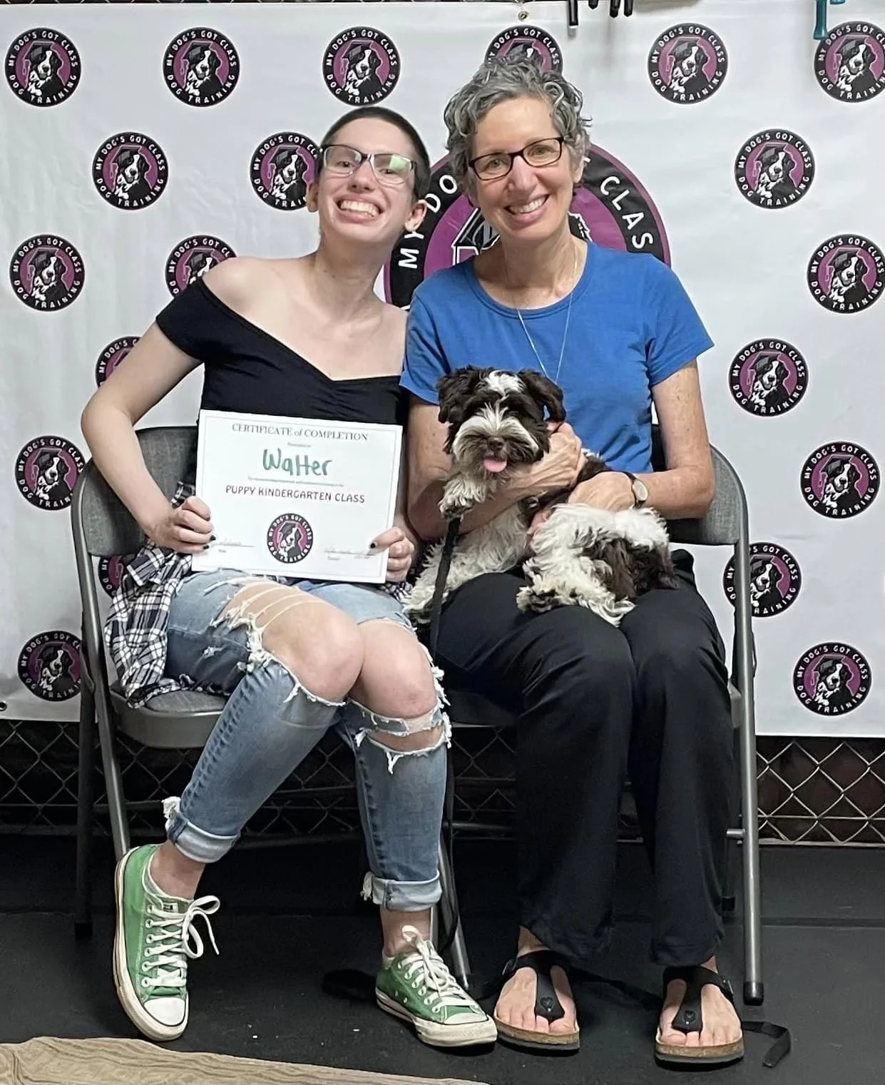 A young woman holding a certificate and smiling, seated next to an older woman with glasses, holding a small puppy, both sitting on gray chairs in front of a backdrop with dog training logos.