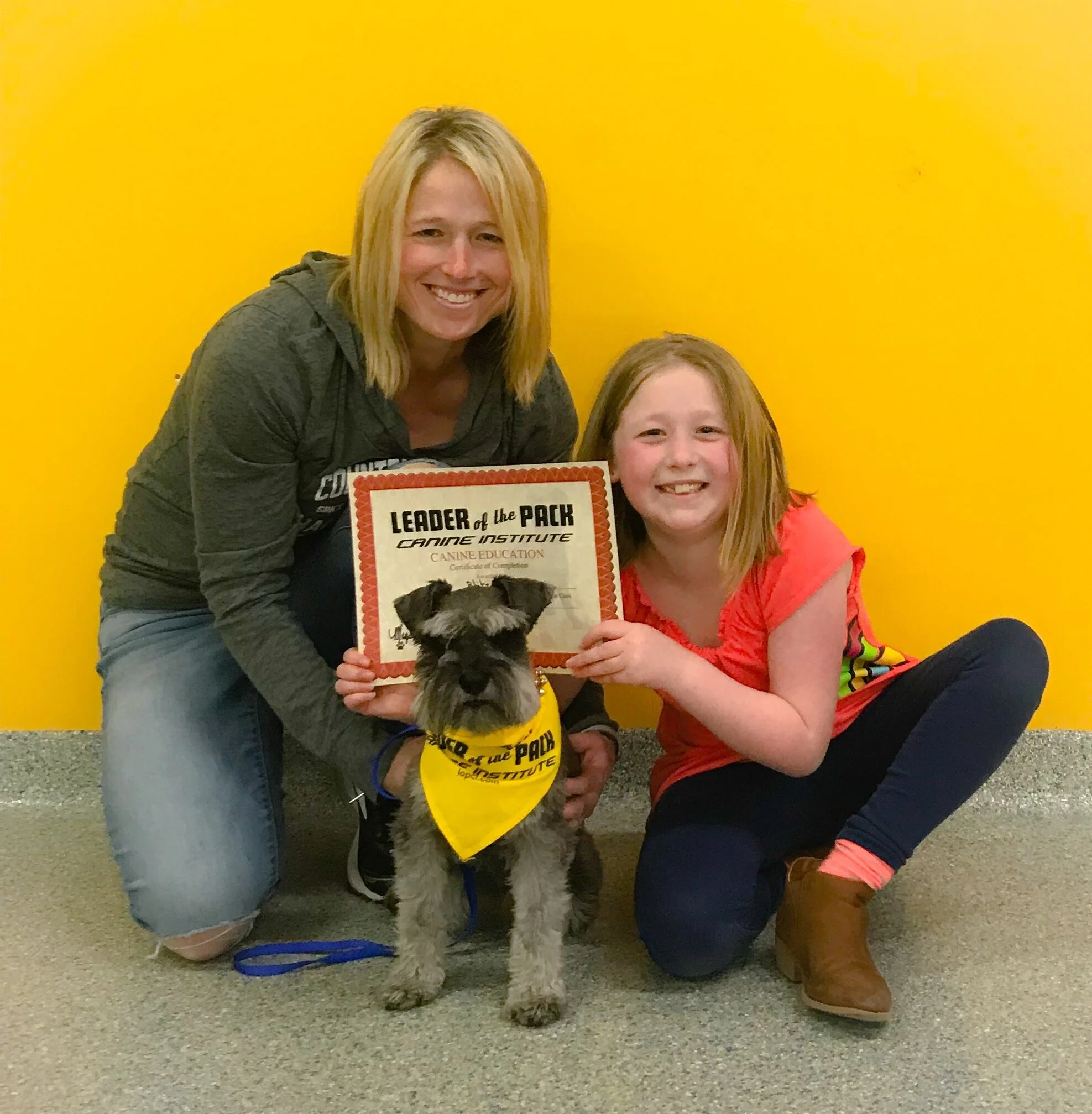 Two women and a young girl smiling indoors against a yellow wall. One woman is holding a certificate, and the girl holds a small dog wearing a yellow bandana that says "LEADER OF THE PACK."