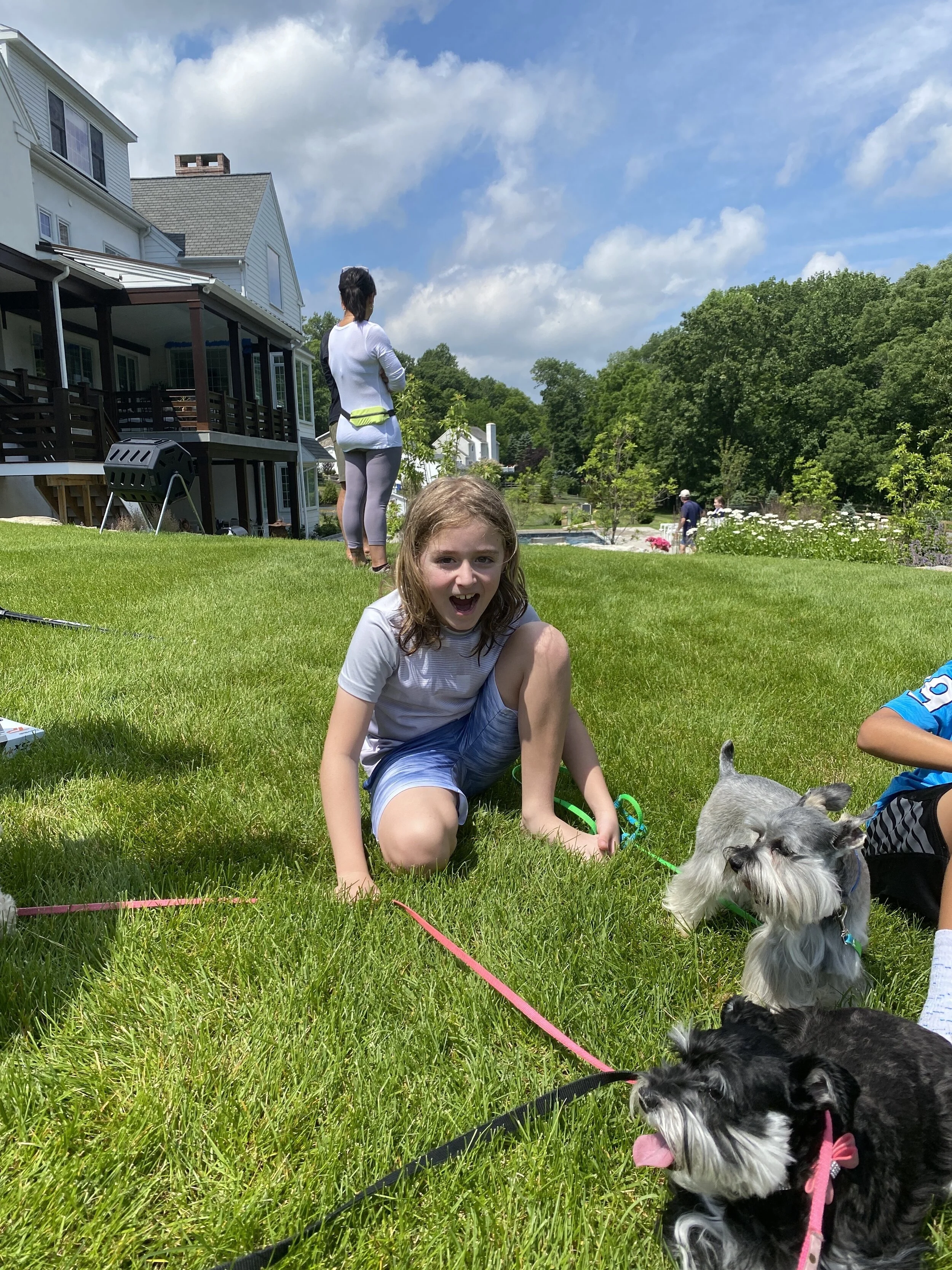 Children and dogs playing on a lush green lawn in a backyard with a white house, trees, and a blue sky with clouds.