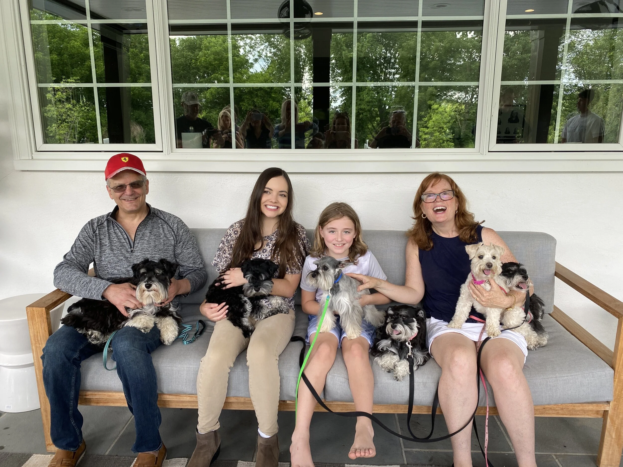 A group of six people and multiple dogs sitting on a gray outdoor sofa in front of a window reflecting several people taking the picture and a lush green outdoor scene.