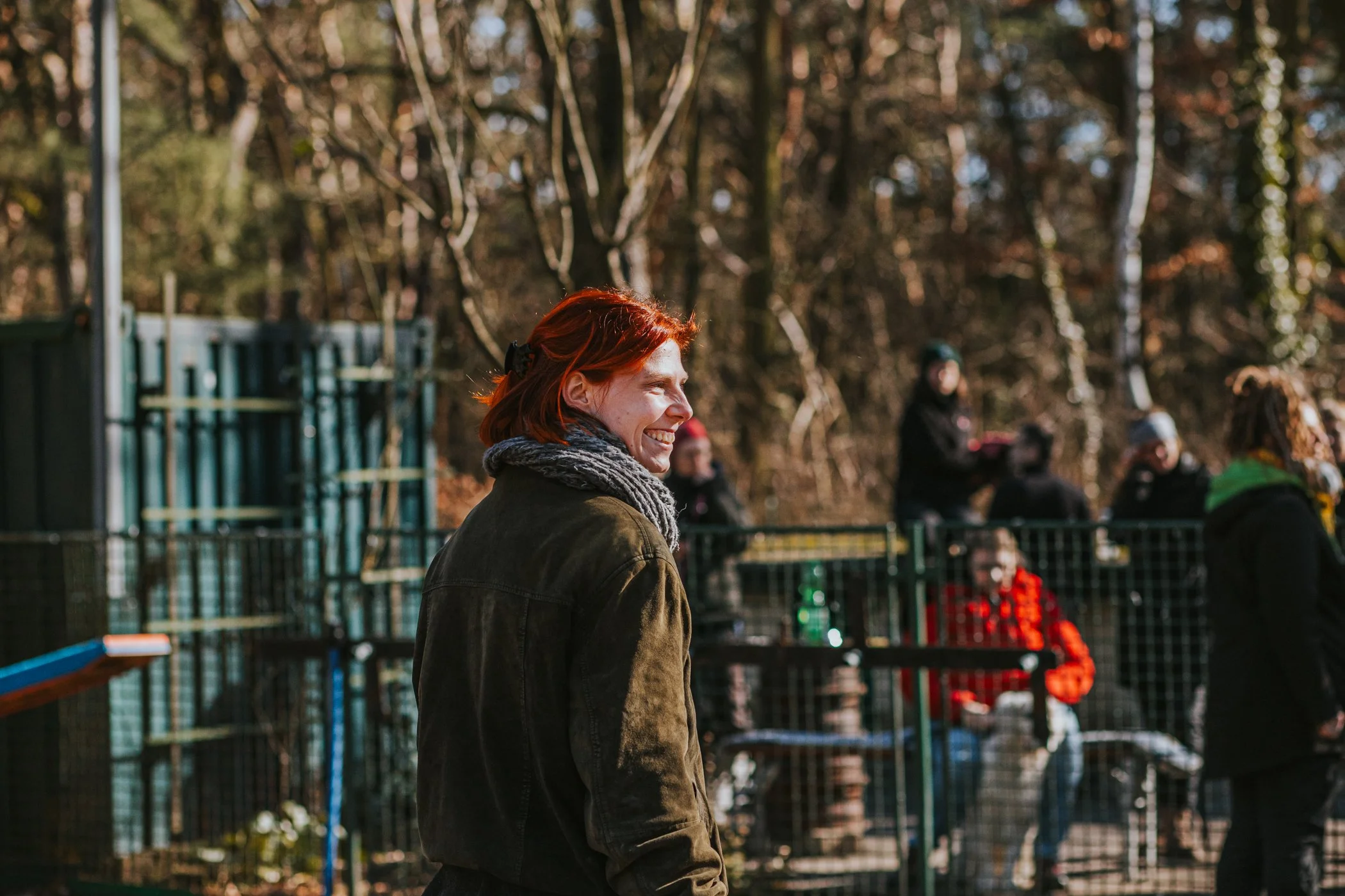 Frau mit roten Haaren und grauem Schal lächelt im Park während eines Outdoor-Events, im Hintergrund Kinder und Erwachsene, unbekannte Aktivitäten.