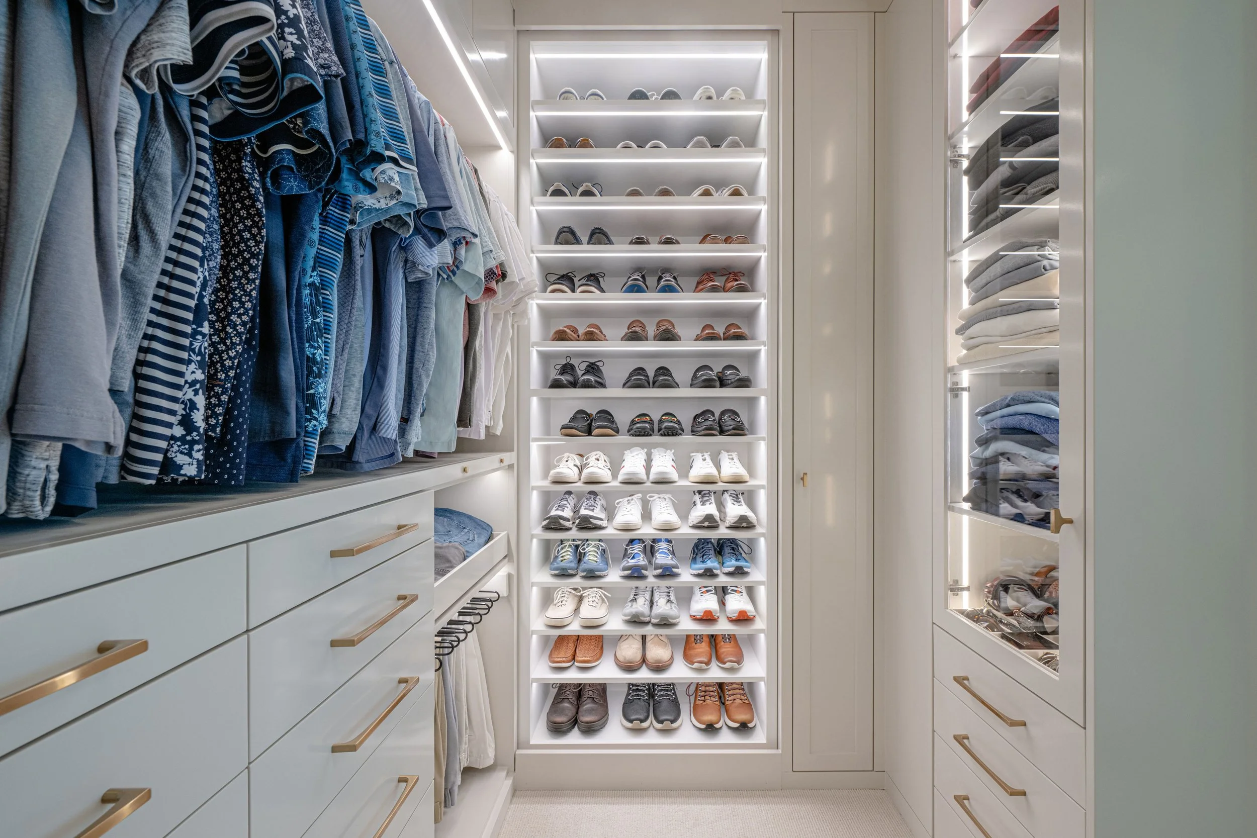 Closet with clothing on the left, shoes on a central illuminated rack, and folded clothes in glass-front cabinets on the right.
