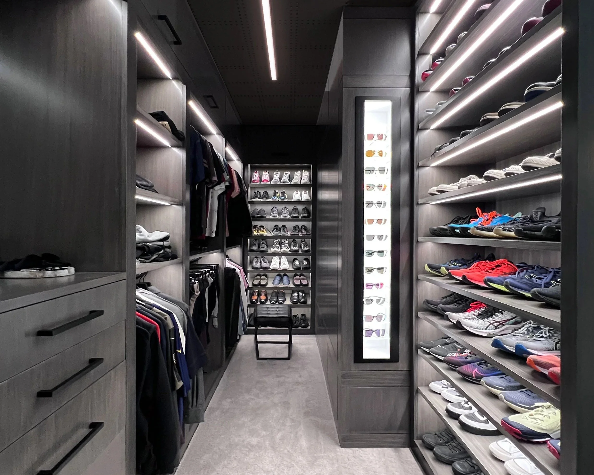 A modern walk-in closet with dark wood shelves and drawers, containing folded clothes, hanging shirts, and a variety of shoes and sunglasses displayed on shelves.