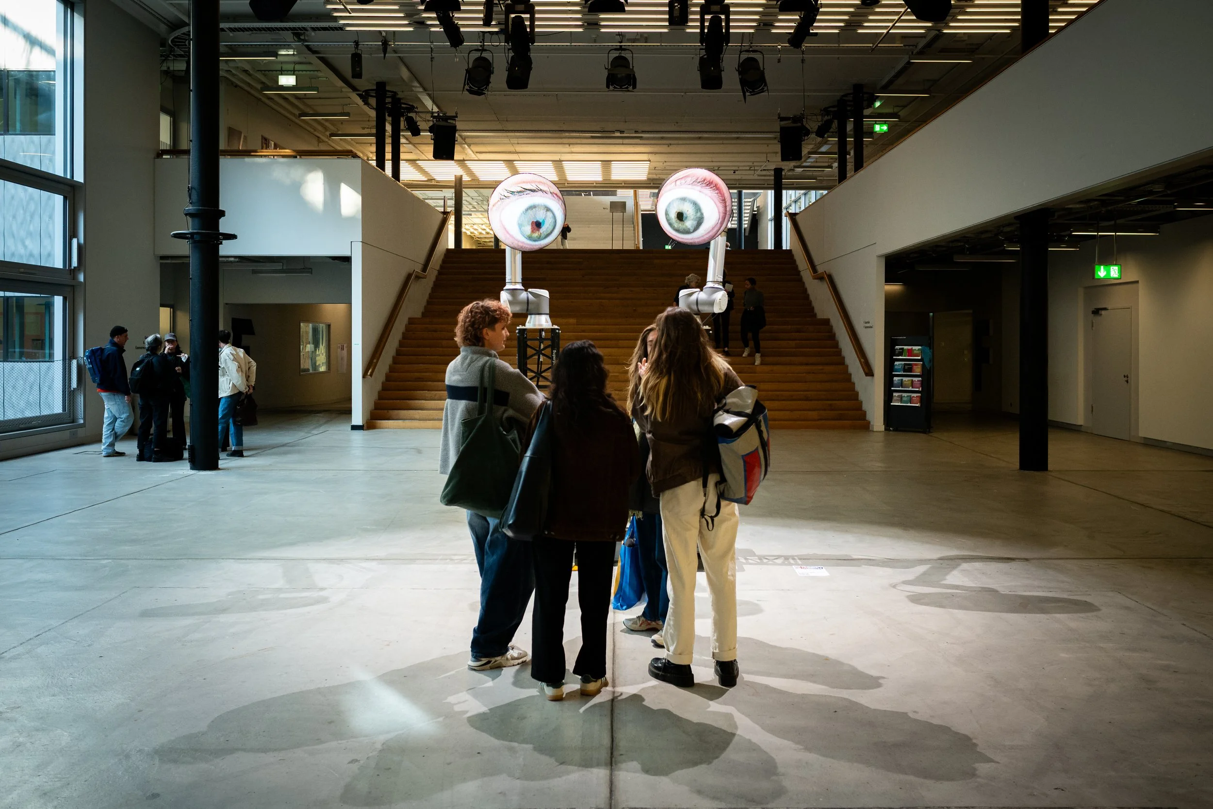Vier Menschen stehen zusammen im Inneren eines Museums oder Ausstellungsraums, schauen nach oben auf eine moderne Kunstinstallation mit zwei großen, schwebenden Augen, die an Robotern befestigt sind. Im Hintergrund sind weitere Besucher, Treppen und 