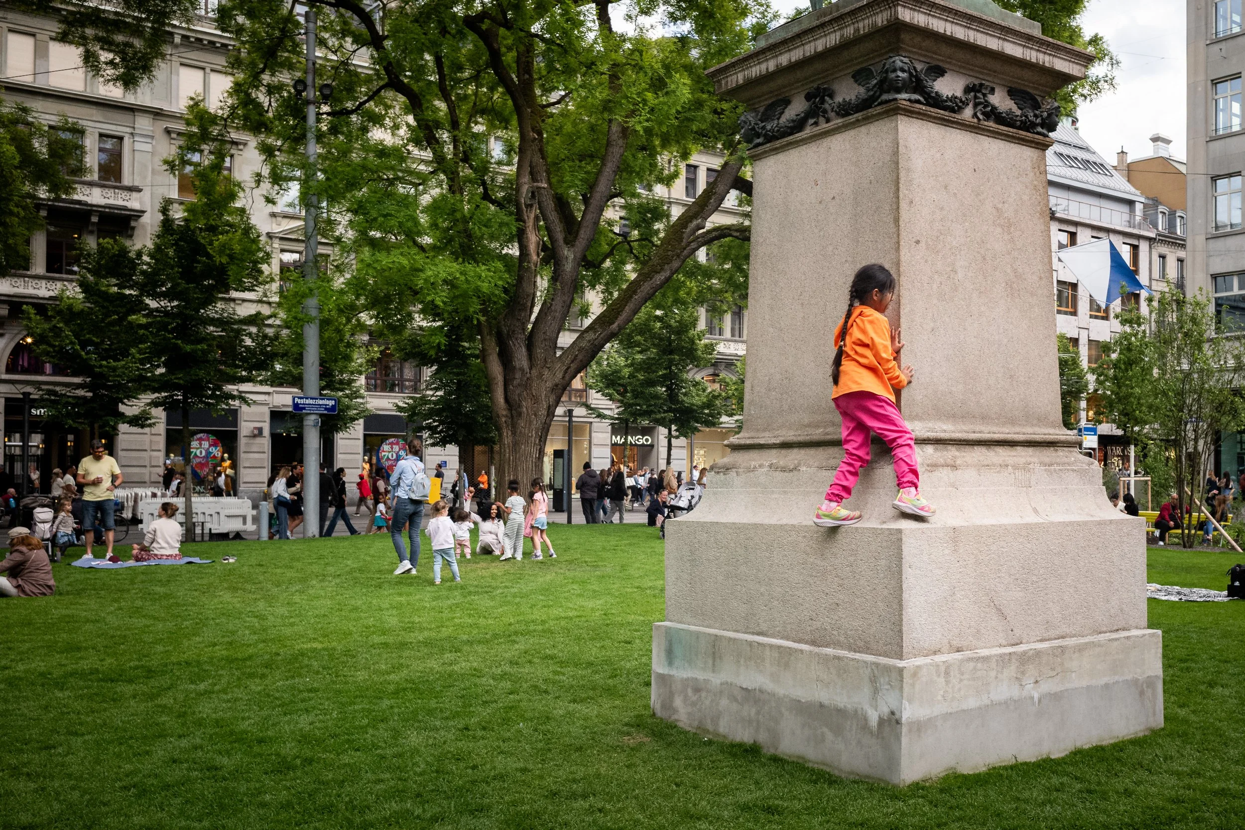 Ein Mädchen in orangefarbener Jacke und pinken Hosen klettert auf eine große Stein-Säule in einem Stadtpark mit grünen Bäumen und mehreren Menschen, die im Hintergrund auf der Wiese sitzen, spazieren gehen oder vor Geschäften stehen.