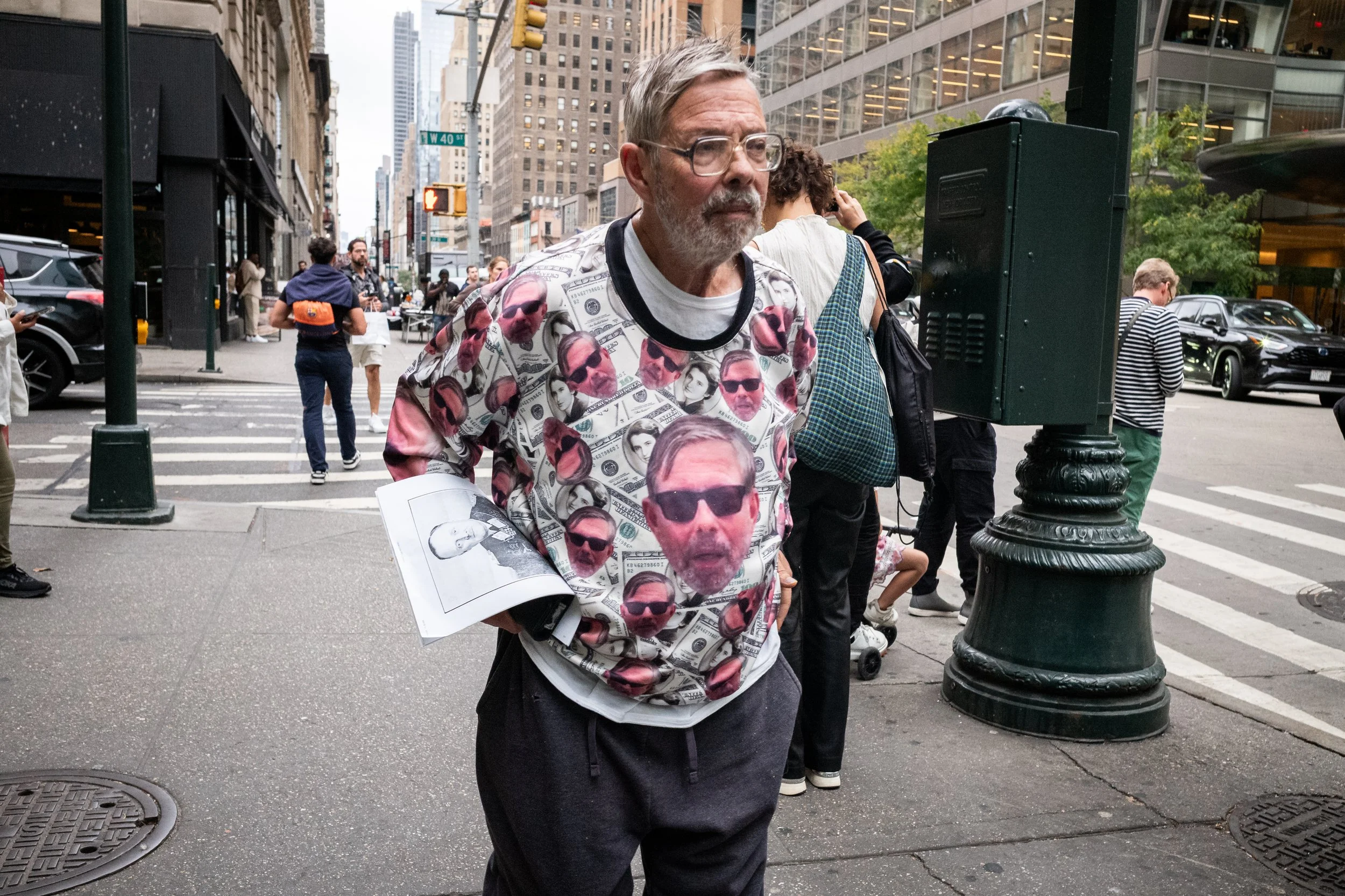 Man in einem Sweatshirt mit Gesichtern im Dollar-Design steht auf einer Stadtstraßenkreuzung.