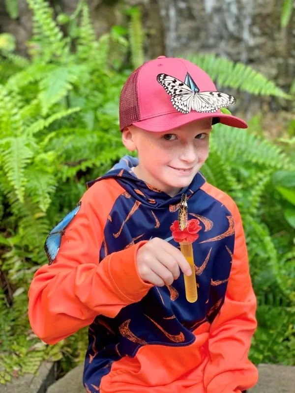 A shy boy smiling as he holds a nectar tube with a butterfly on it and has a butterfly on his red cap