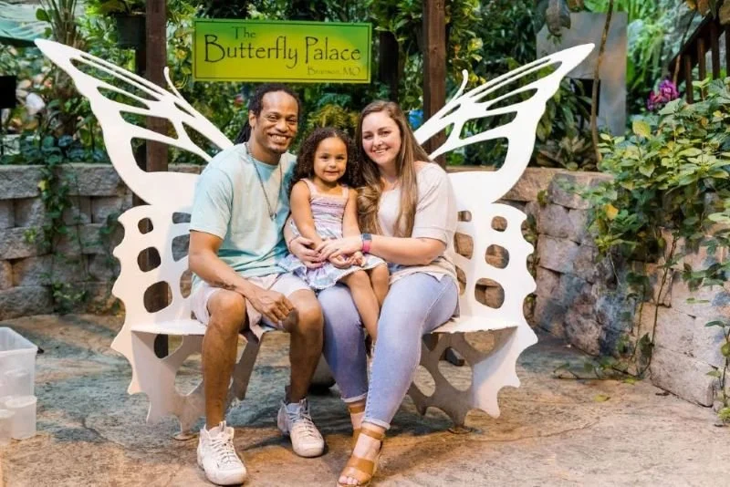 father and mother holding young daughter in their laps, all sitting on a metal bench shaped like a butterfly