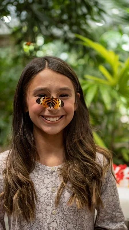 A smiling woman with a butterfly perched on her nose, surrounded by green foliage in a lush, indoor garden or greenhouse.