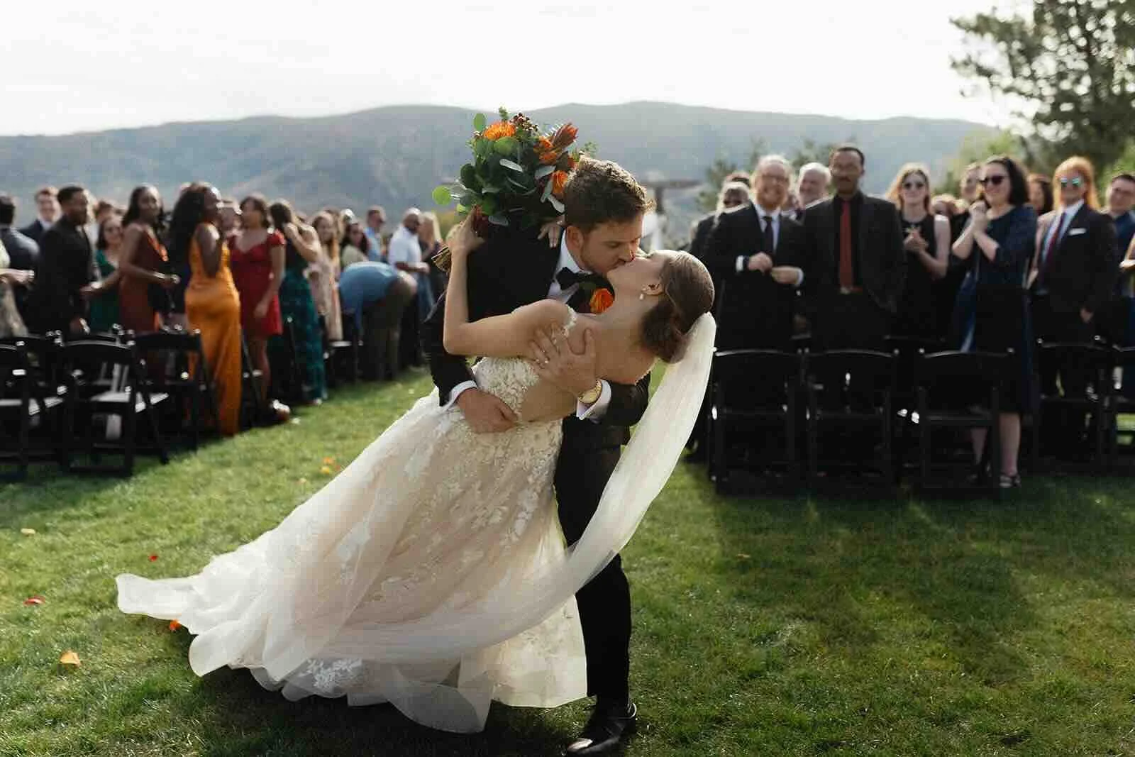 Wedding couple kissing at the end of the aisle and dipping in front of their family and friends. There are Utah mountains in the background.