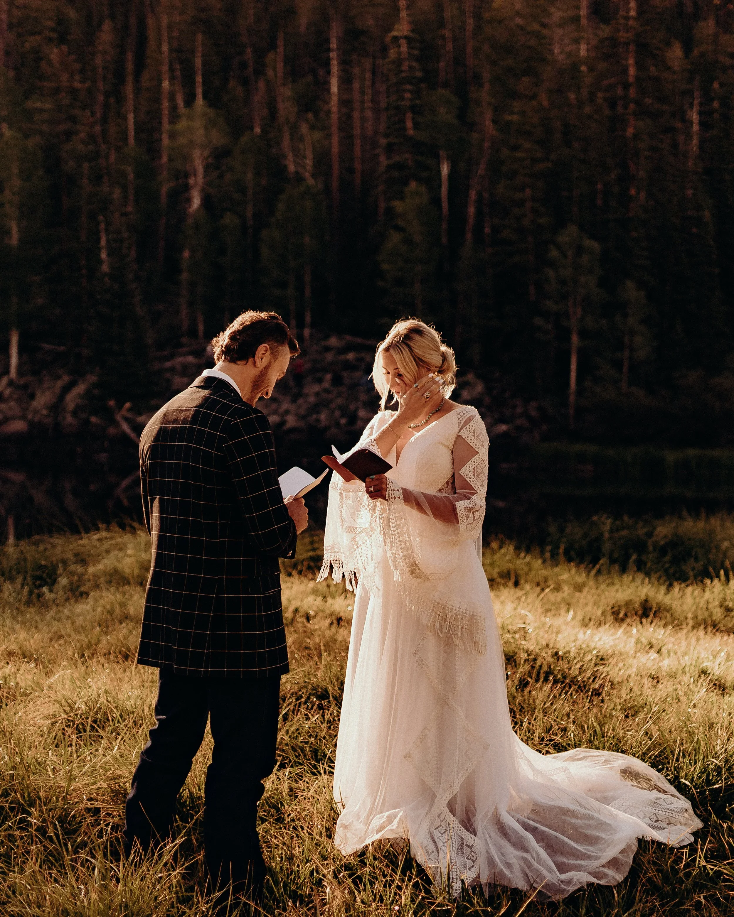 A couple getting married outdoors at sunset, with the officiant holding a book and reading vows, in a grassy area near a lake with trees in the background.