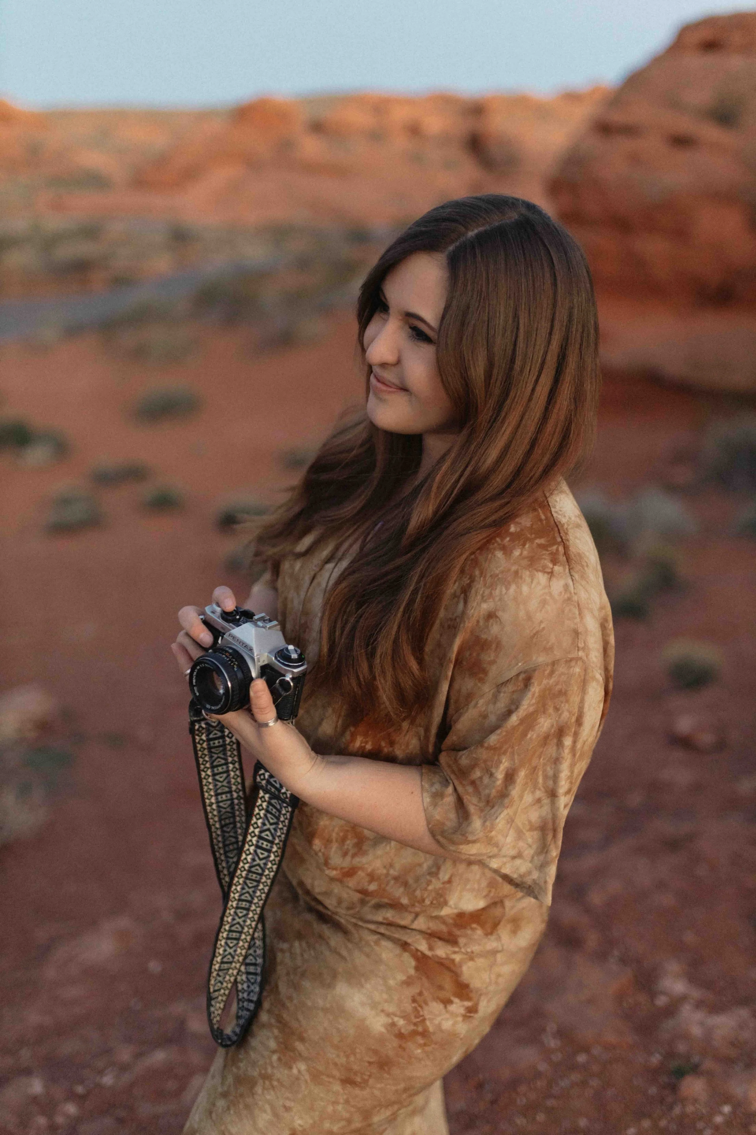 A woman with long brown hair, wearing a brown tie-dye shirt, holding a vintage camera with a patterned strap, standing in a desert landscape with red rocks.