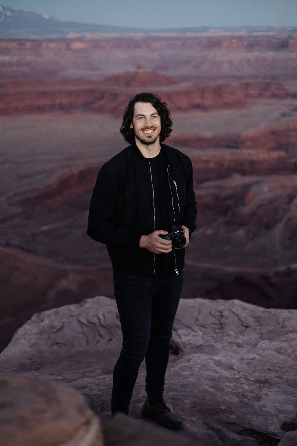 A man with dark, curly hair and a beard, wearing a black jacket and jeans, stands on a rocky ledge holding a camera, with the Grand Canyon in the background during dusk.