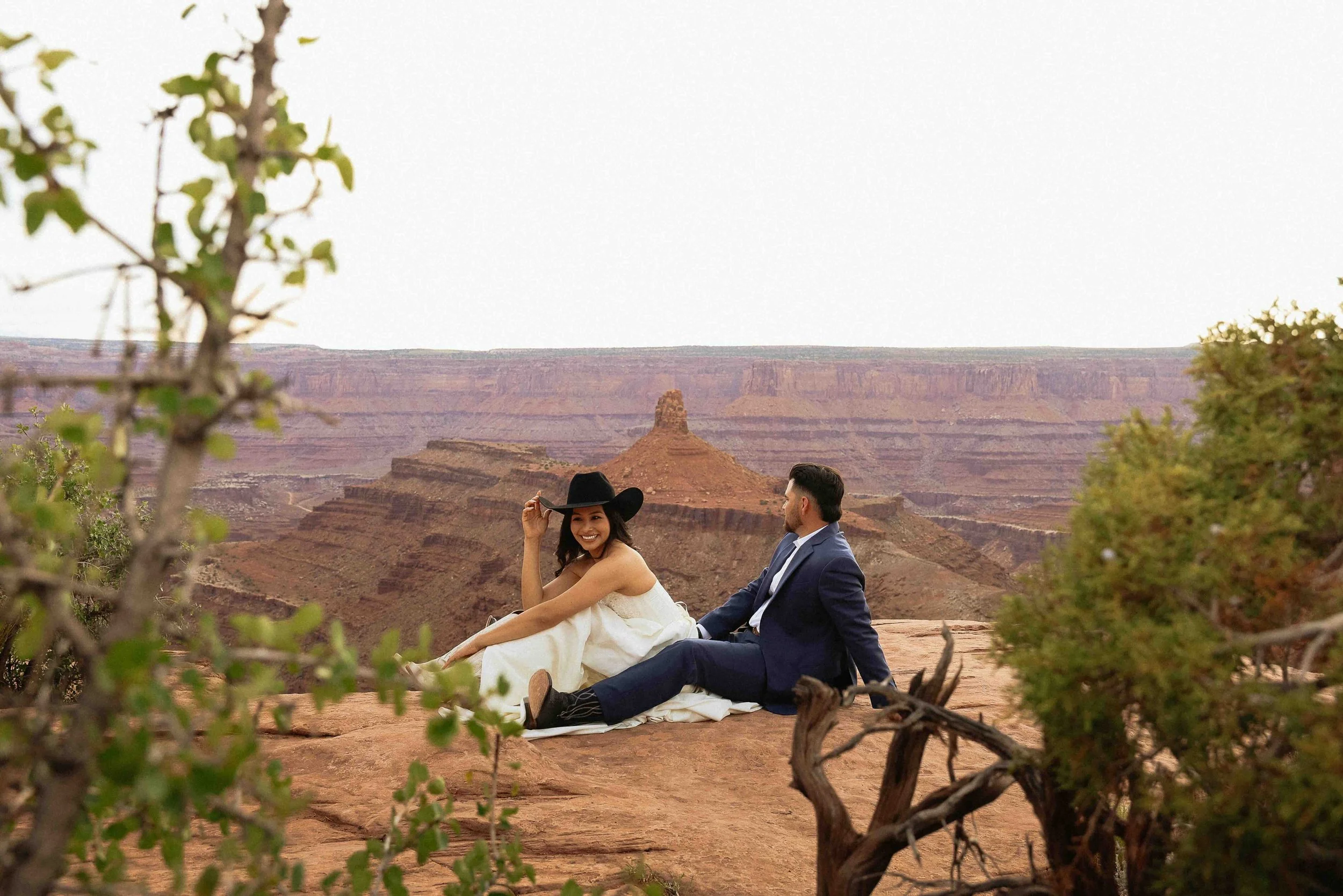 Bride and groom sitting on a cliffside of Moab, UT. The bride is wearing the groom's cowboy hat and laughing. Plants are in the foreground.