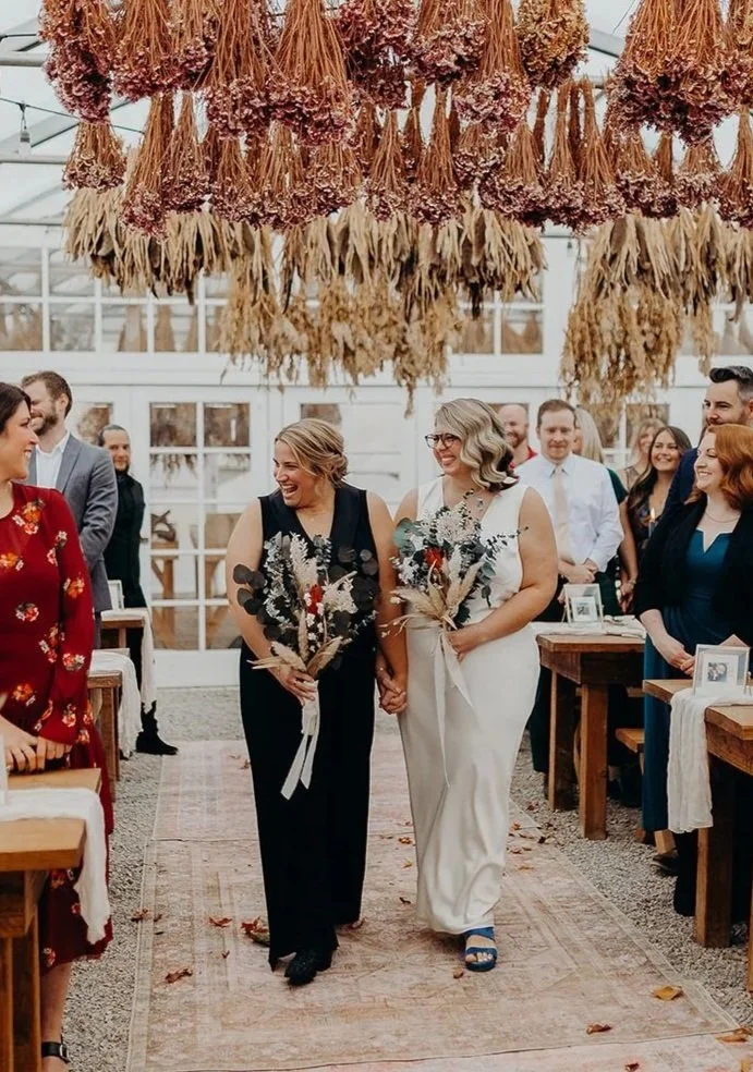 Two brides walking down the aisle together in a white garden house.
