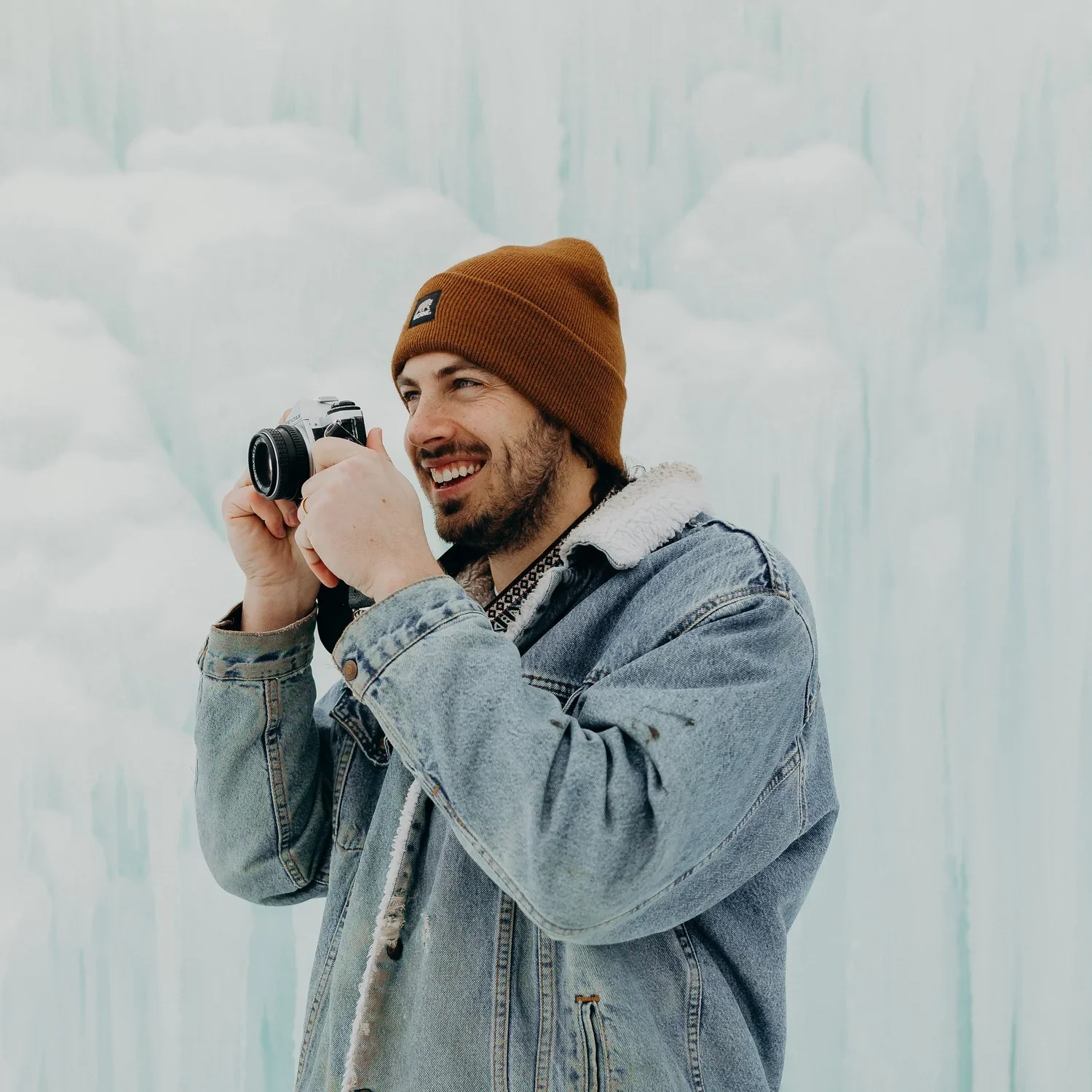 A man wearing a brown beanie and denim jacket holding a camera and smiling, standing in front of an icy blue glacier.