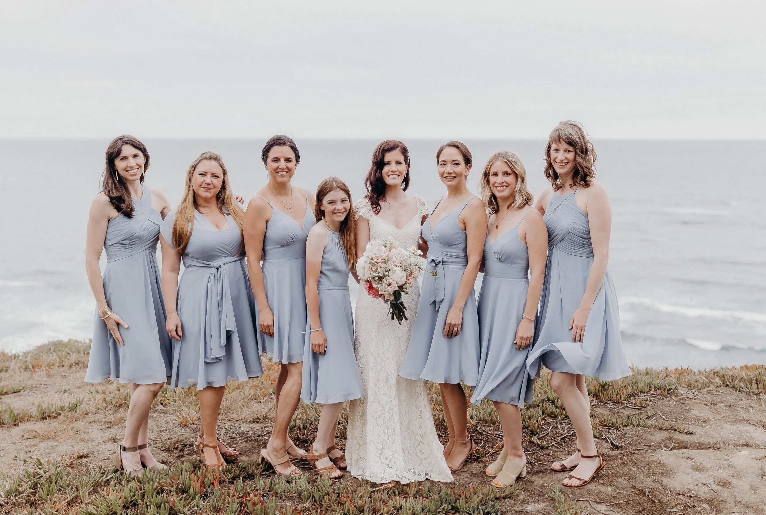 Bridesmaids in blue standing in front of the ocean with the bride