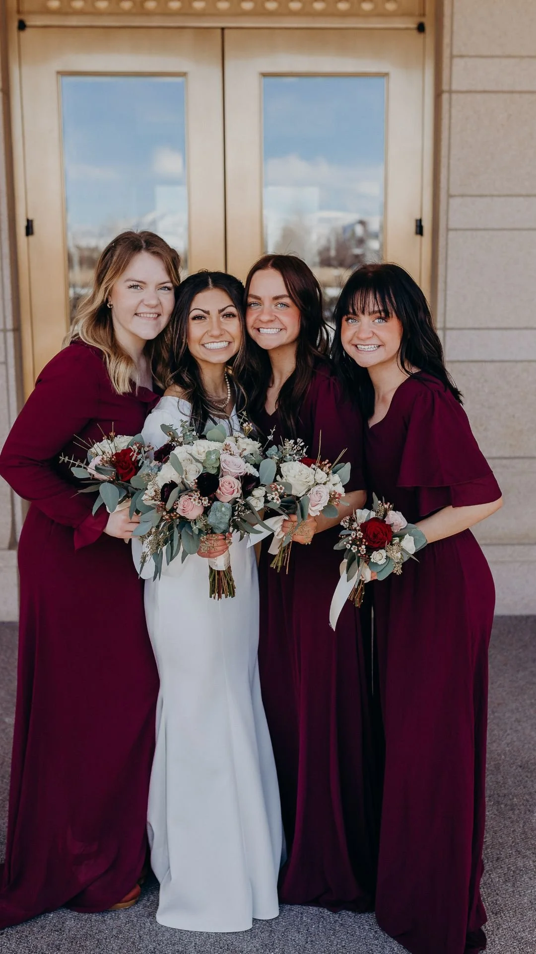 Bridesmaids in red, holding their bouquets with the bride in front of gold doors