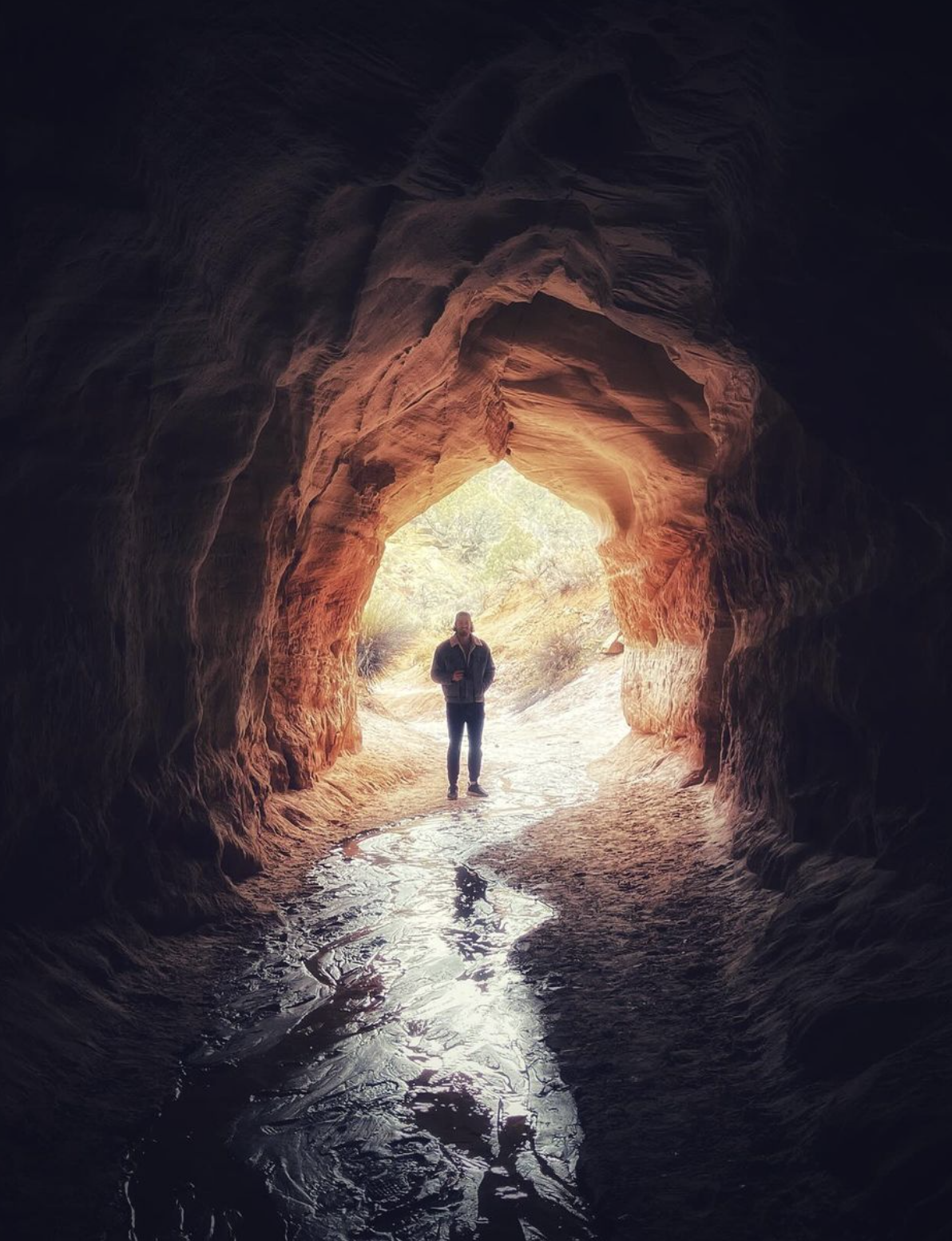 A person standing at the exit of a cave with a river running out of it
