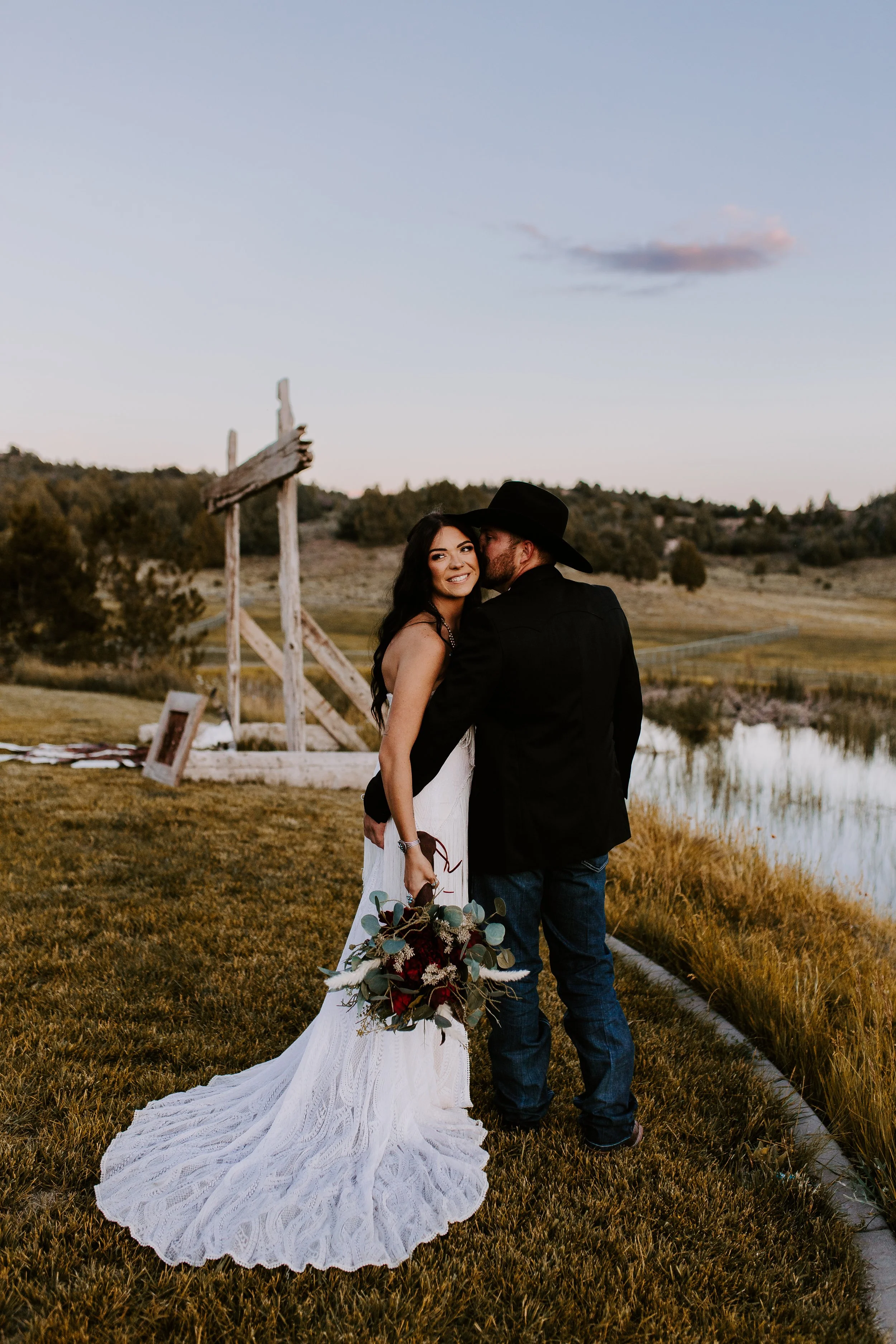 A bride holding flowers smiling at the camera while the groom hugs her
