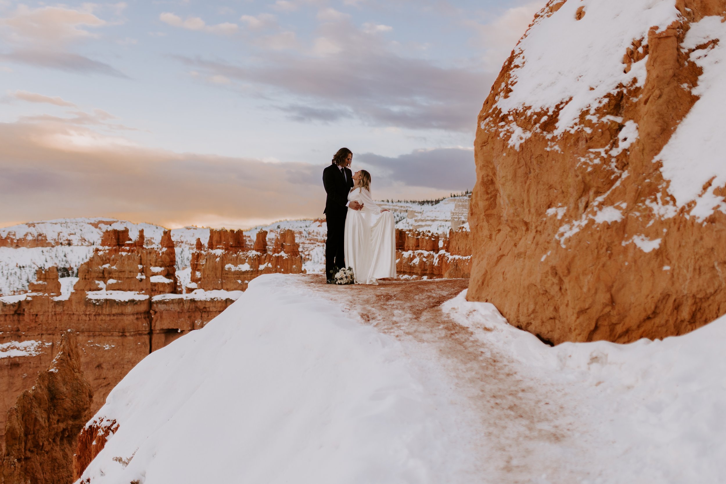 A bride and groom standing on a snowy trail with red rock in the background