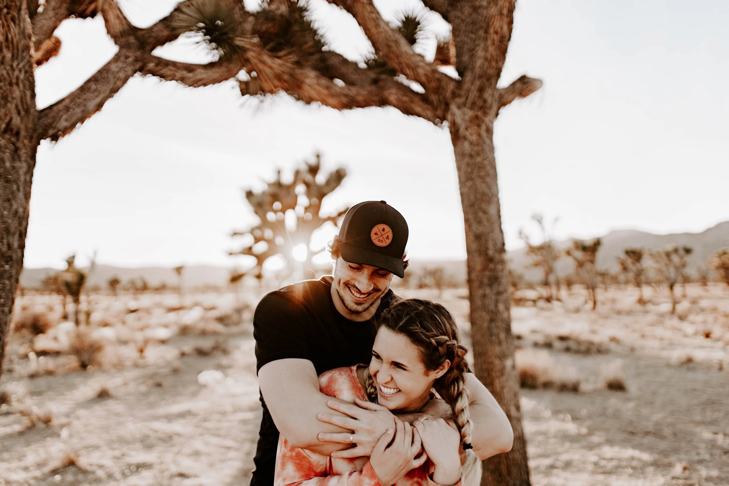 Couple hugging each other and laughing in front of desert with sun setting behind them