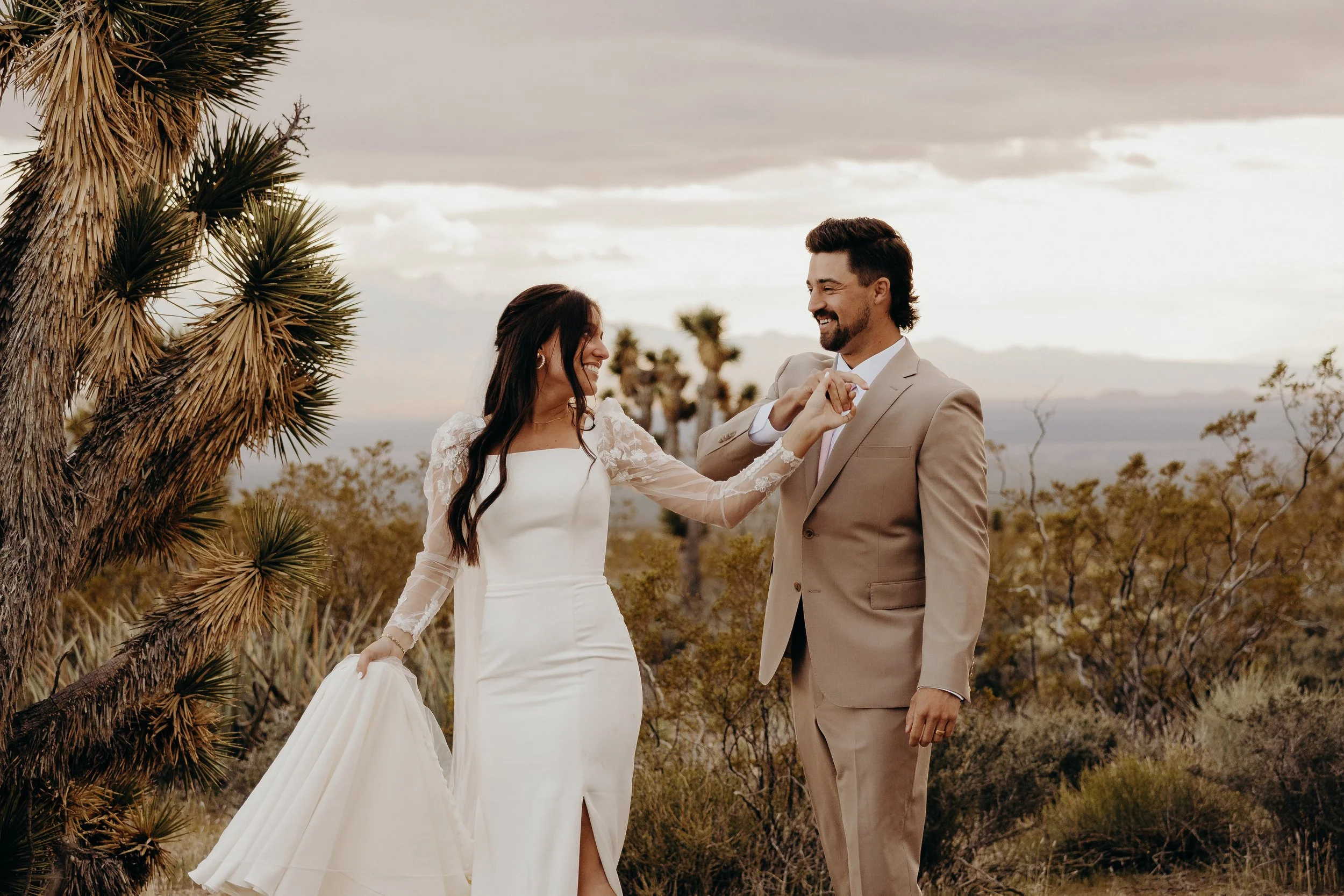 Bride and groom holding hands and twirling in desert with joshua trees surrounding them and mountains in the distance
