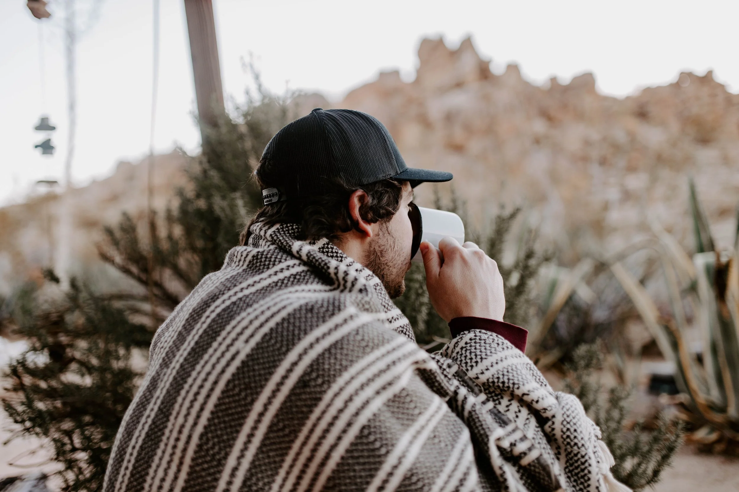 Man in desert blanket drinking coffee in front of boulders and desert plants