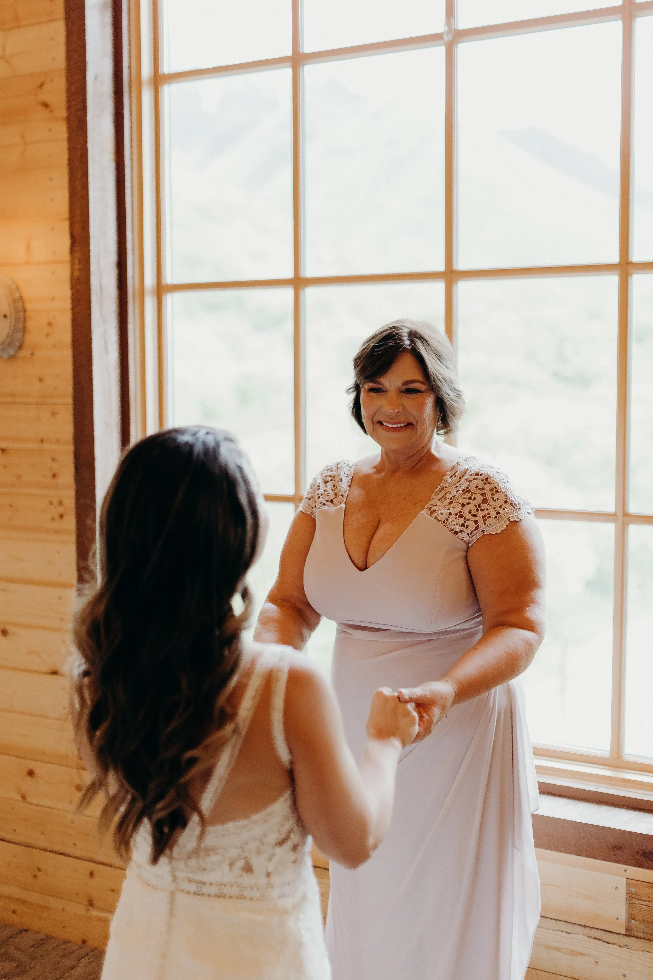 Mother of bride seeing bride for the first time and smiling in front of large window of barn