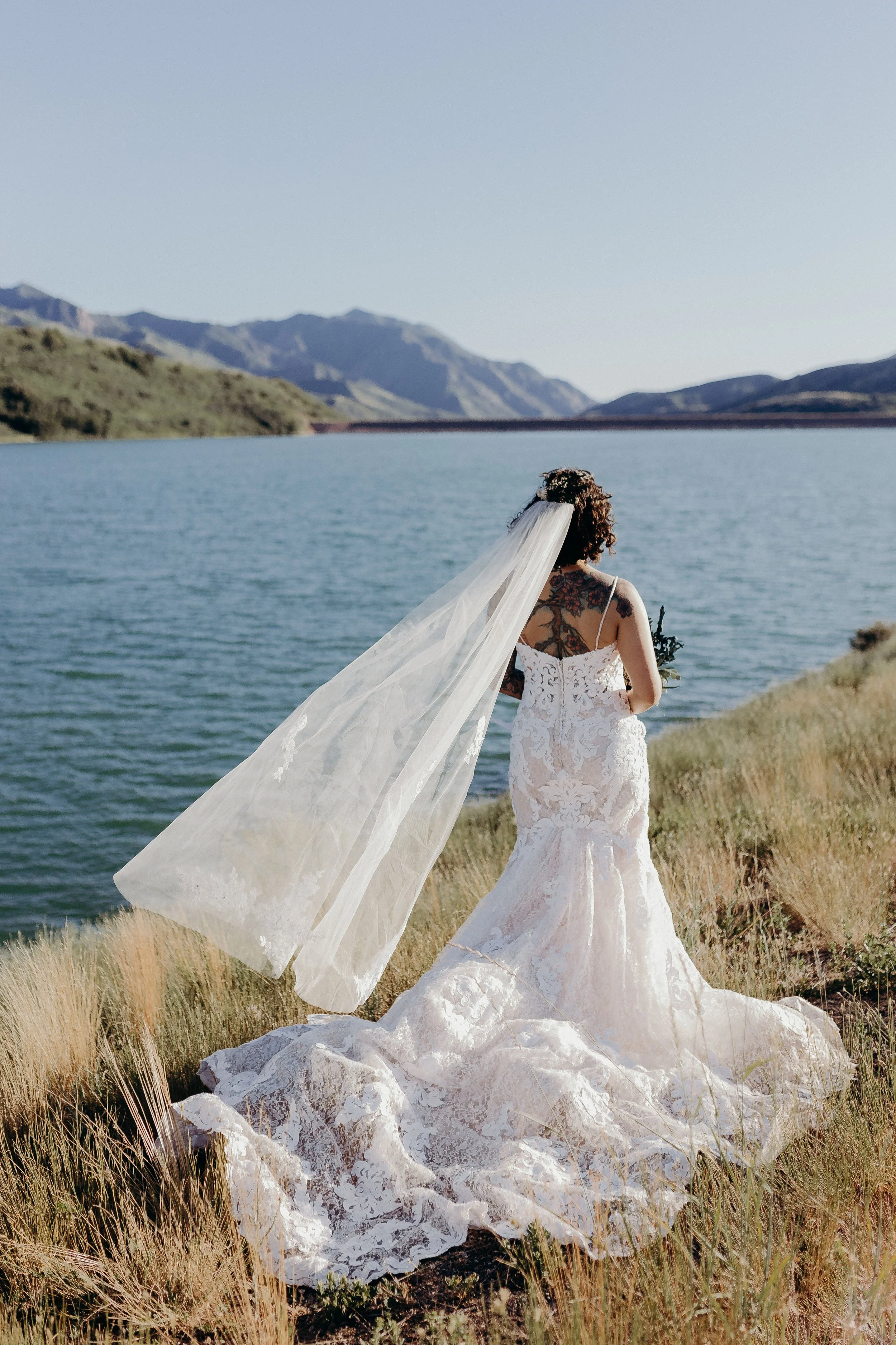 Bride looking at reservoir with mountains in the background. She has tattoos on her back and her veil is blowing back. 