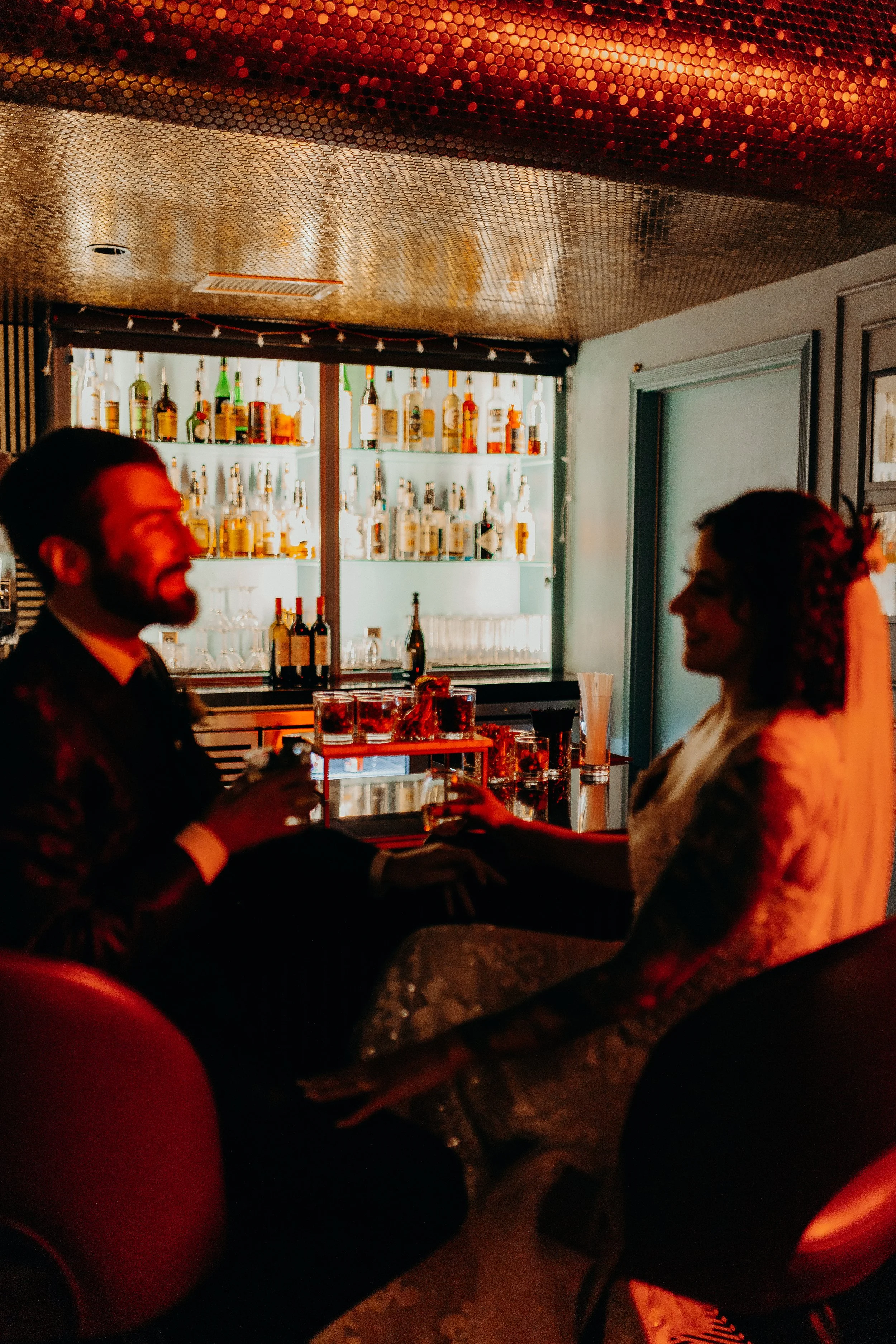 Red bar lights on bride and groom smiling at each other with drinks in front of bar