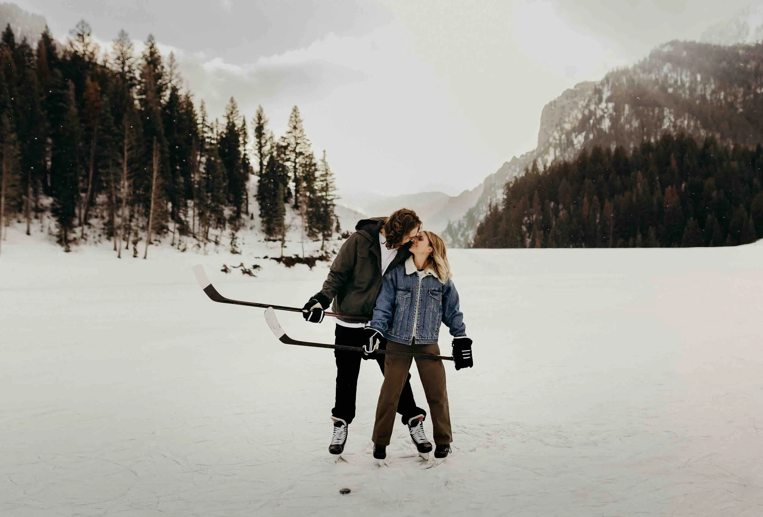 Hockey couple holding onto their hockey sticks on a frozen lake with mountains in the background.