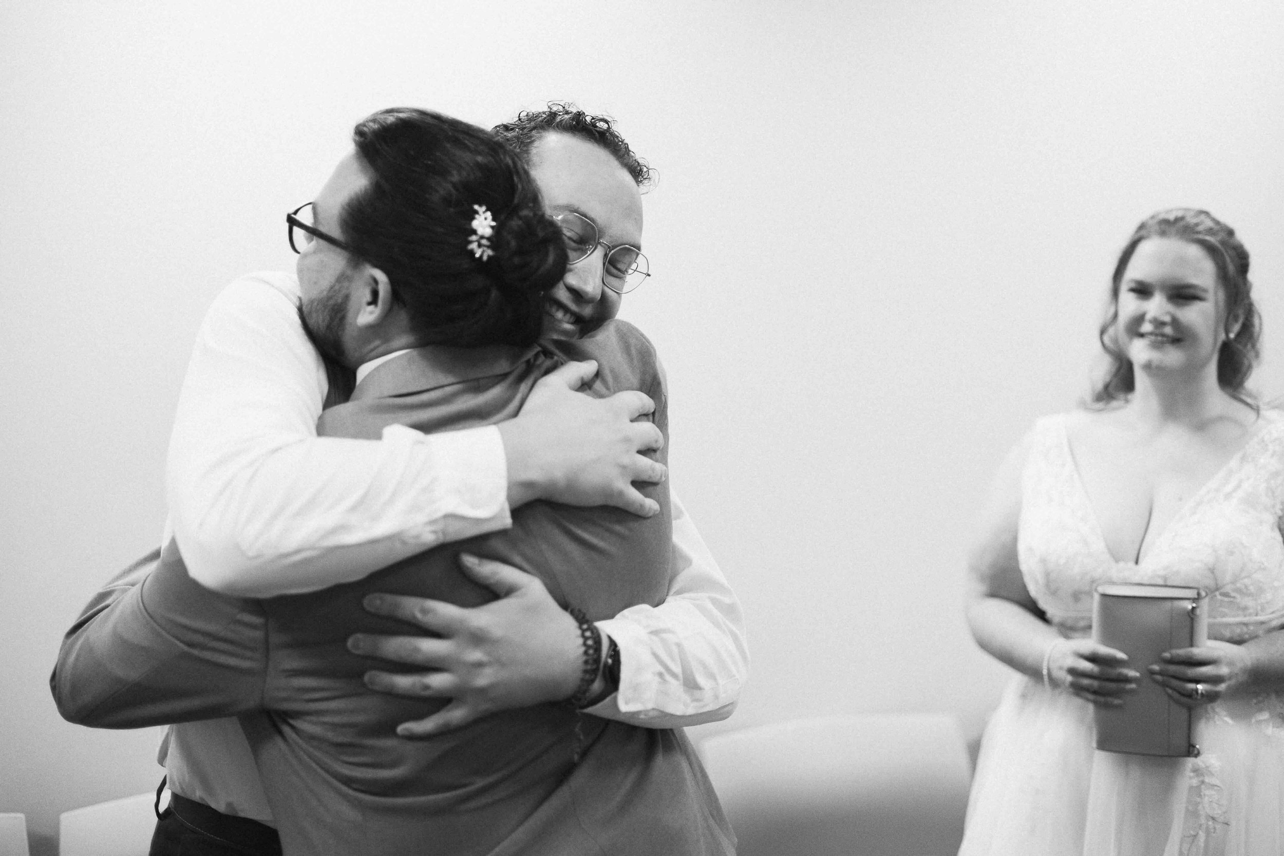Groom hugging his witness/friend, with bride smiling at them in the background.