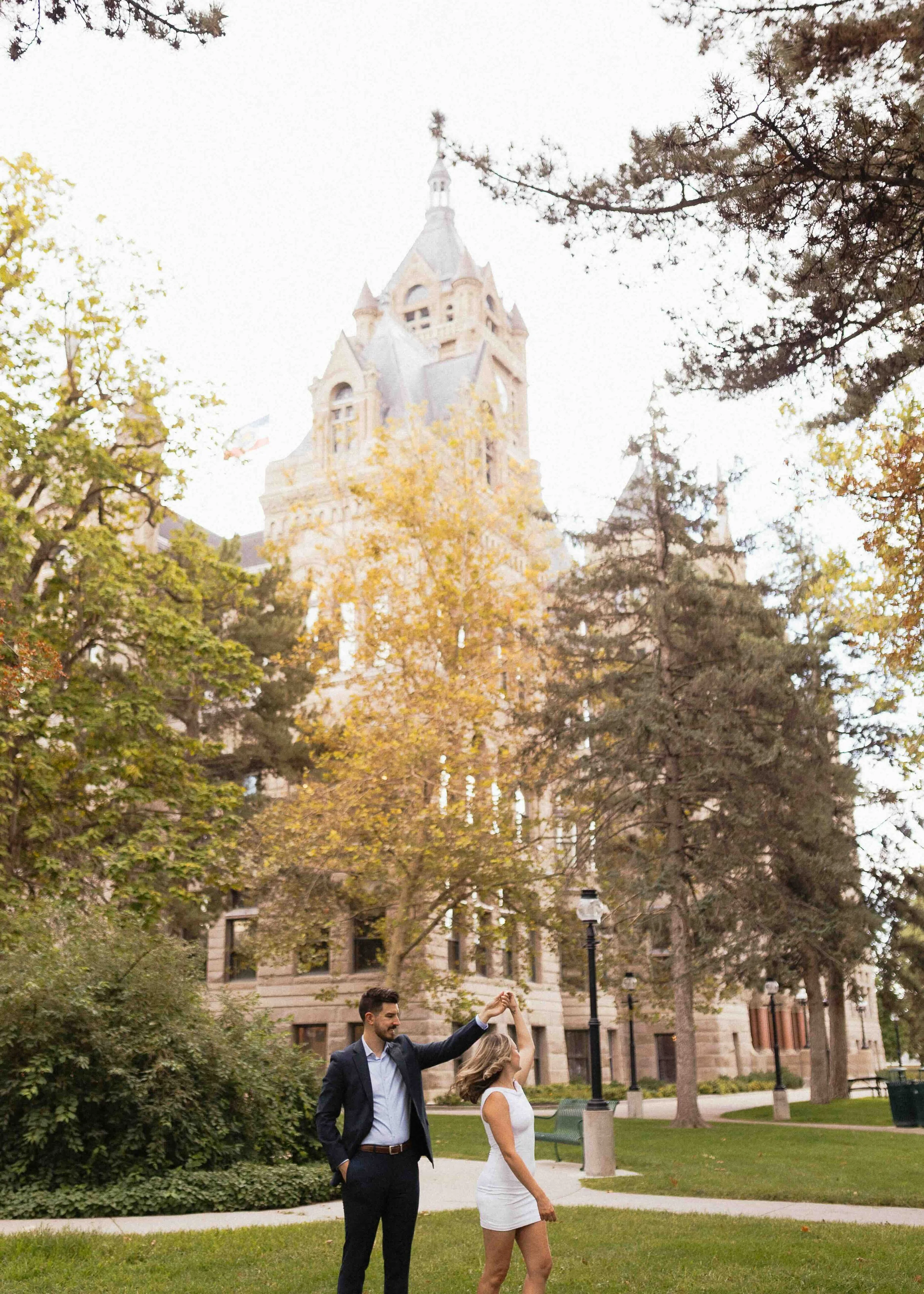 Groom twirling bride in front of SLC City hall with trees where the leaves are starting to change colors.