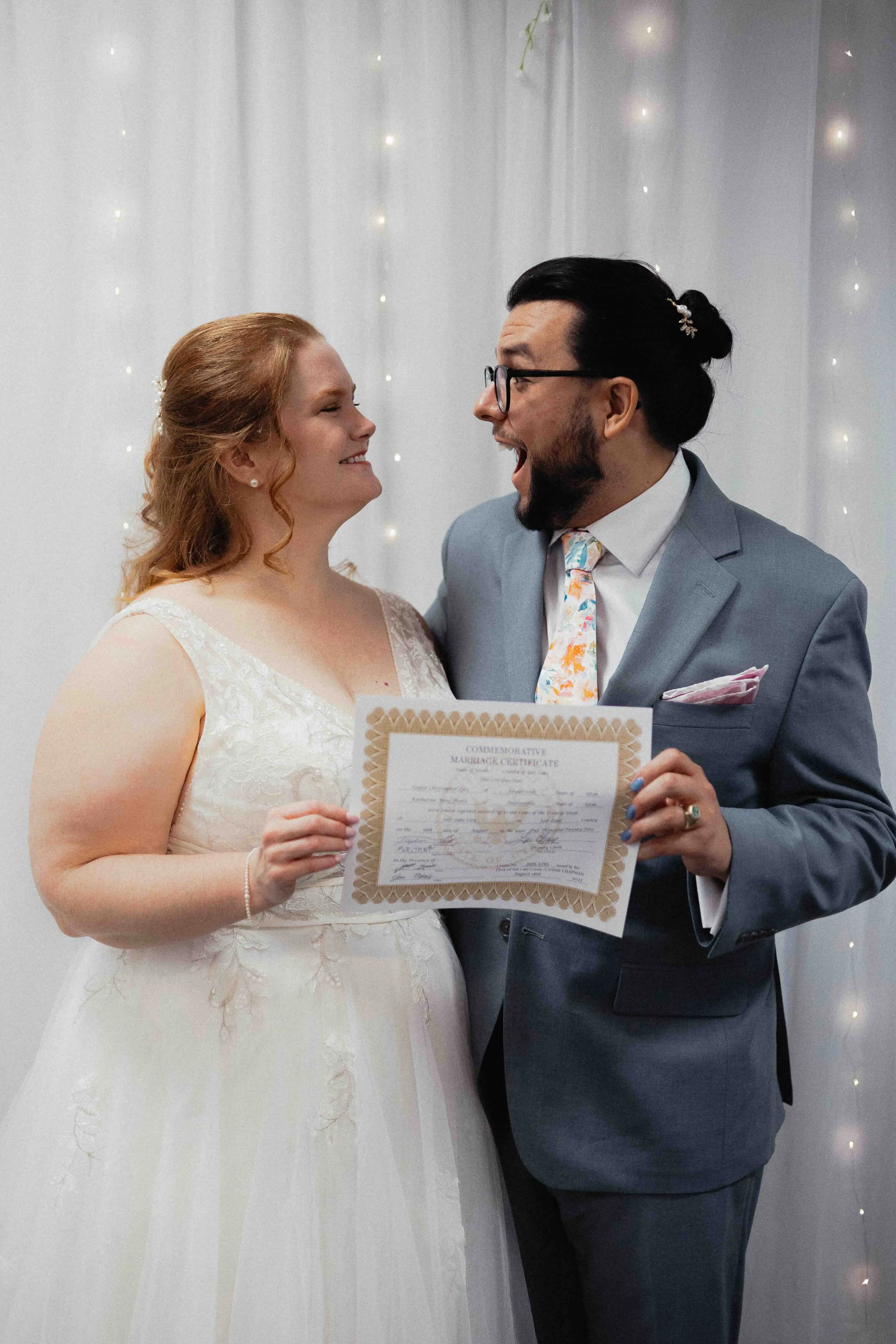 Bride and groom holding marriage certificate in front of white curtain with lights.