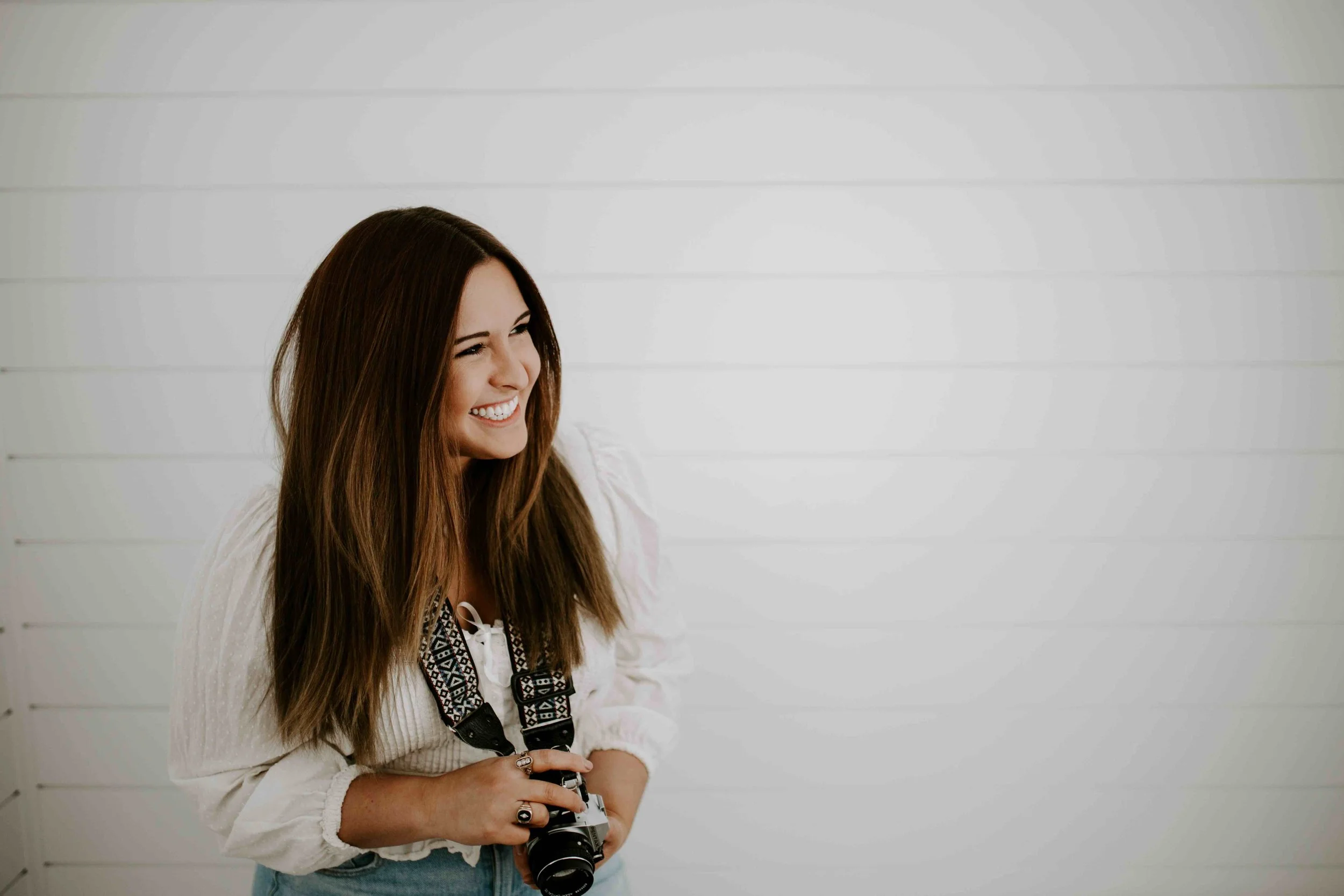 A smiling young woman with long brown hair holding a camera, standing against a white wooden wall.