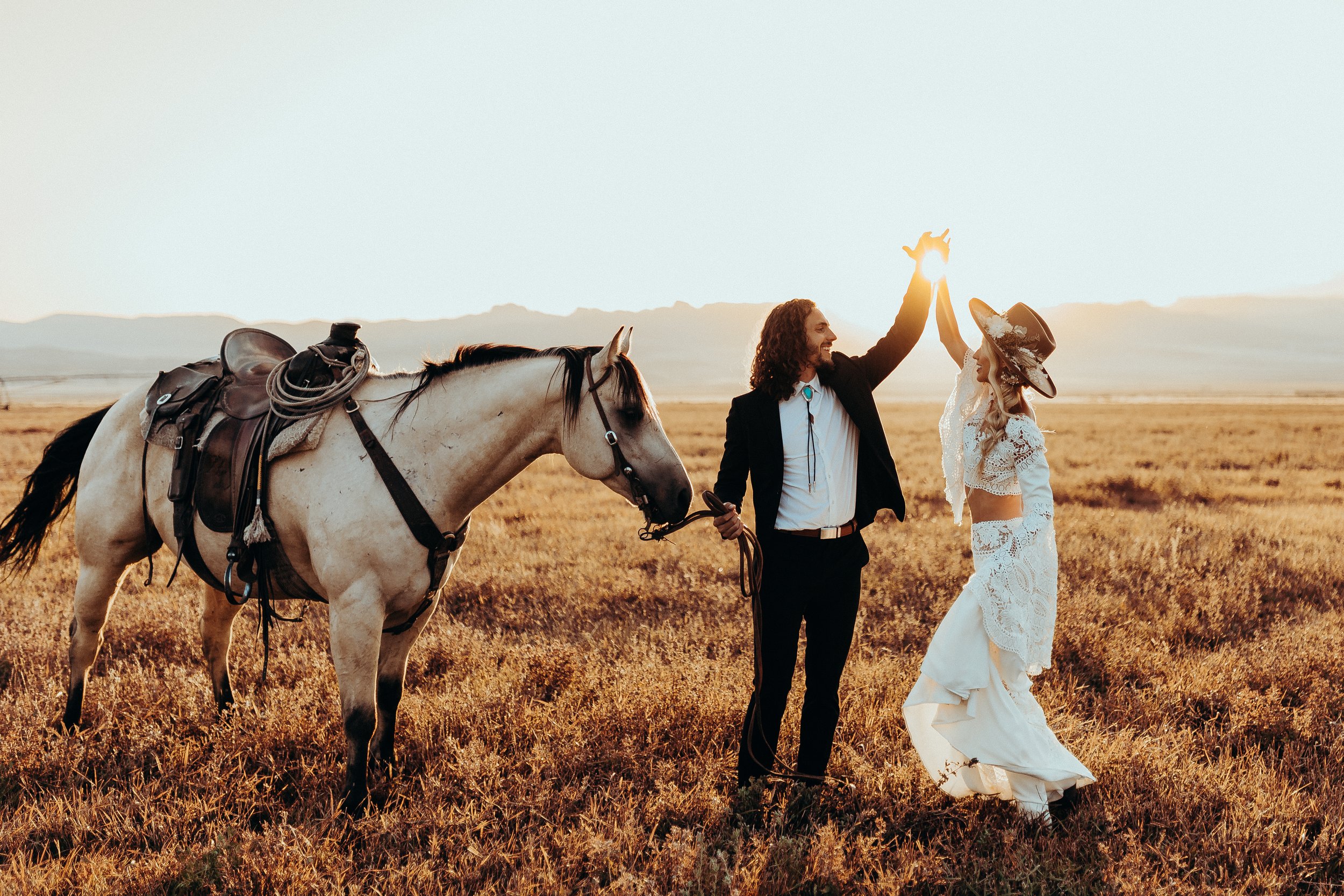 A man in a suit holding hands with a woman in a white dress, with a horse, in a yellow grass field