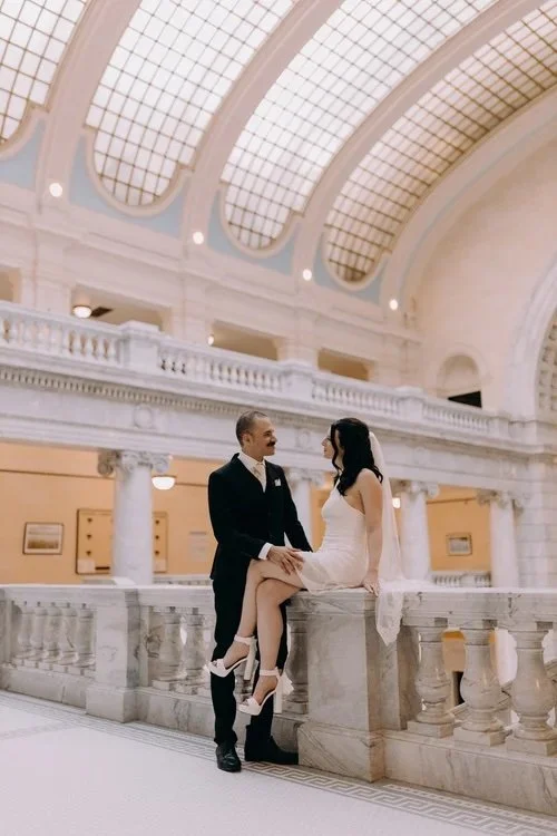 Bride sitting on a marbled railing by her groom with a curved windowed ceiling in the background.