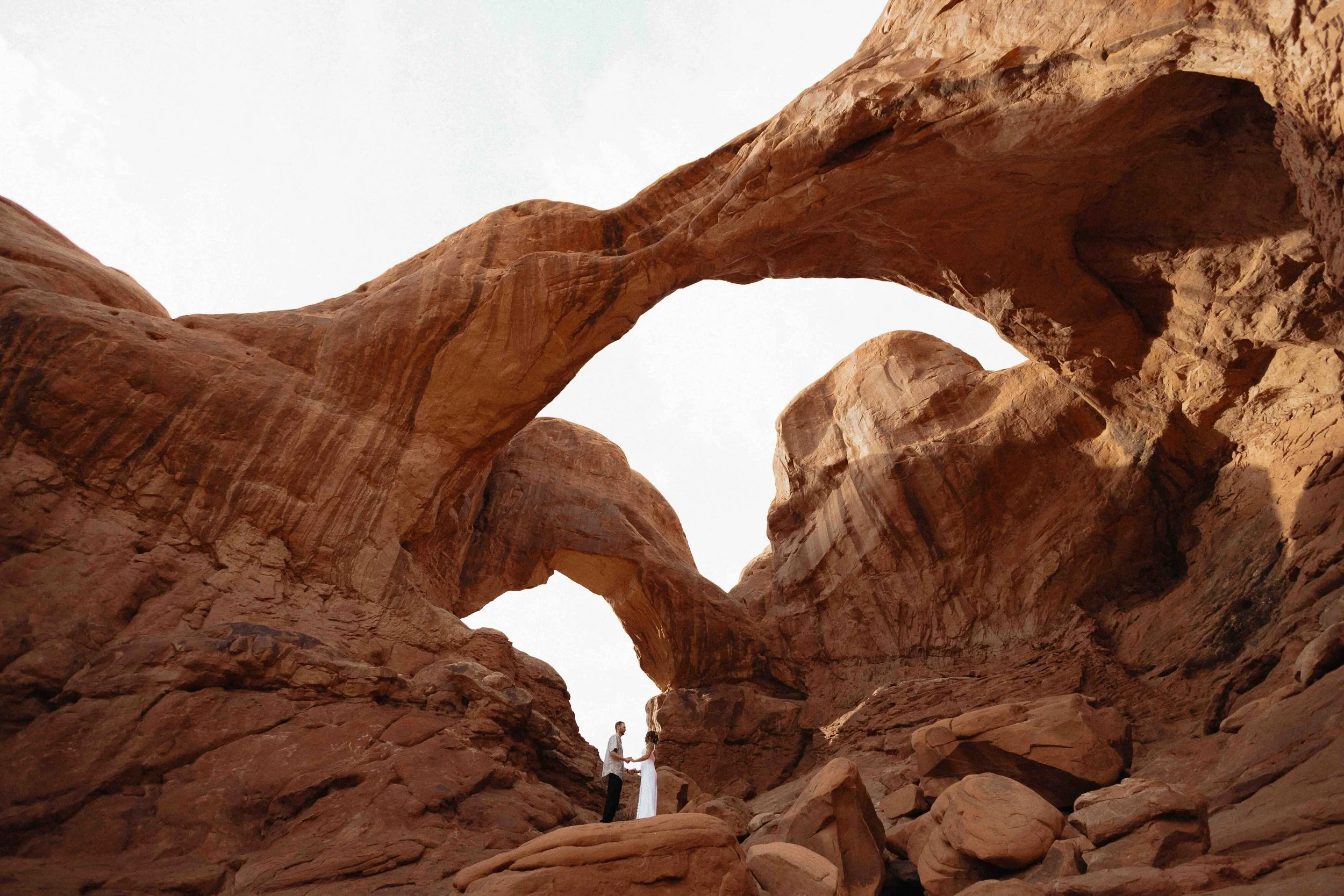 Bride and groom standing far away inside of two red rock arches in Moab, called the Double Arch.