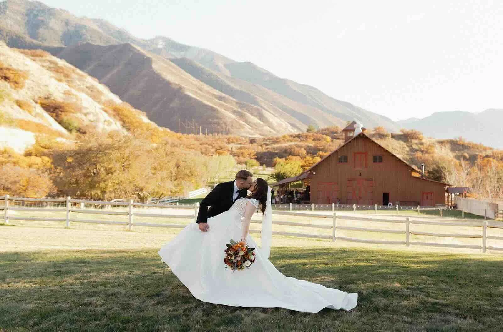 Wedding couple with bouquet dipping in front of the mountain views and a brown barn in the background in Utah.