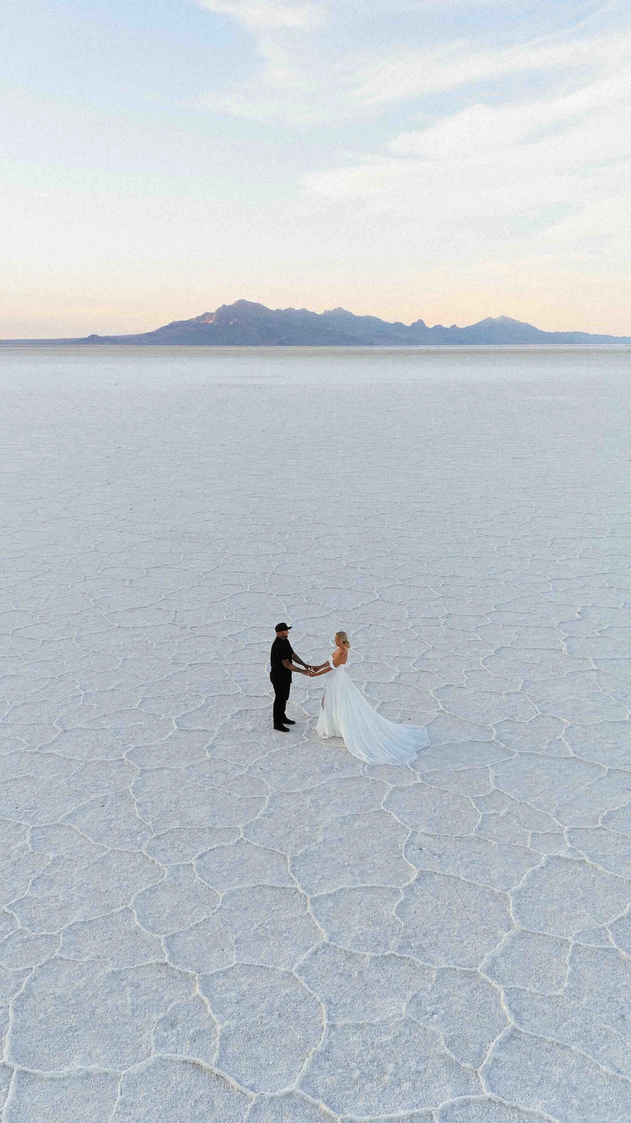 Groom and bride holding hands on the Bonneville salt flats with mountains in the background.