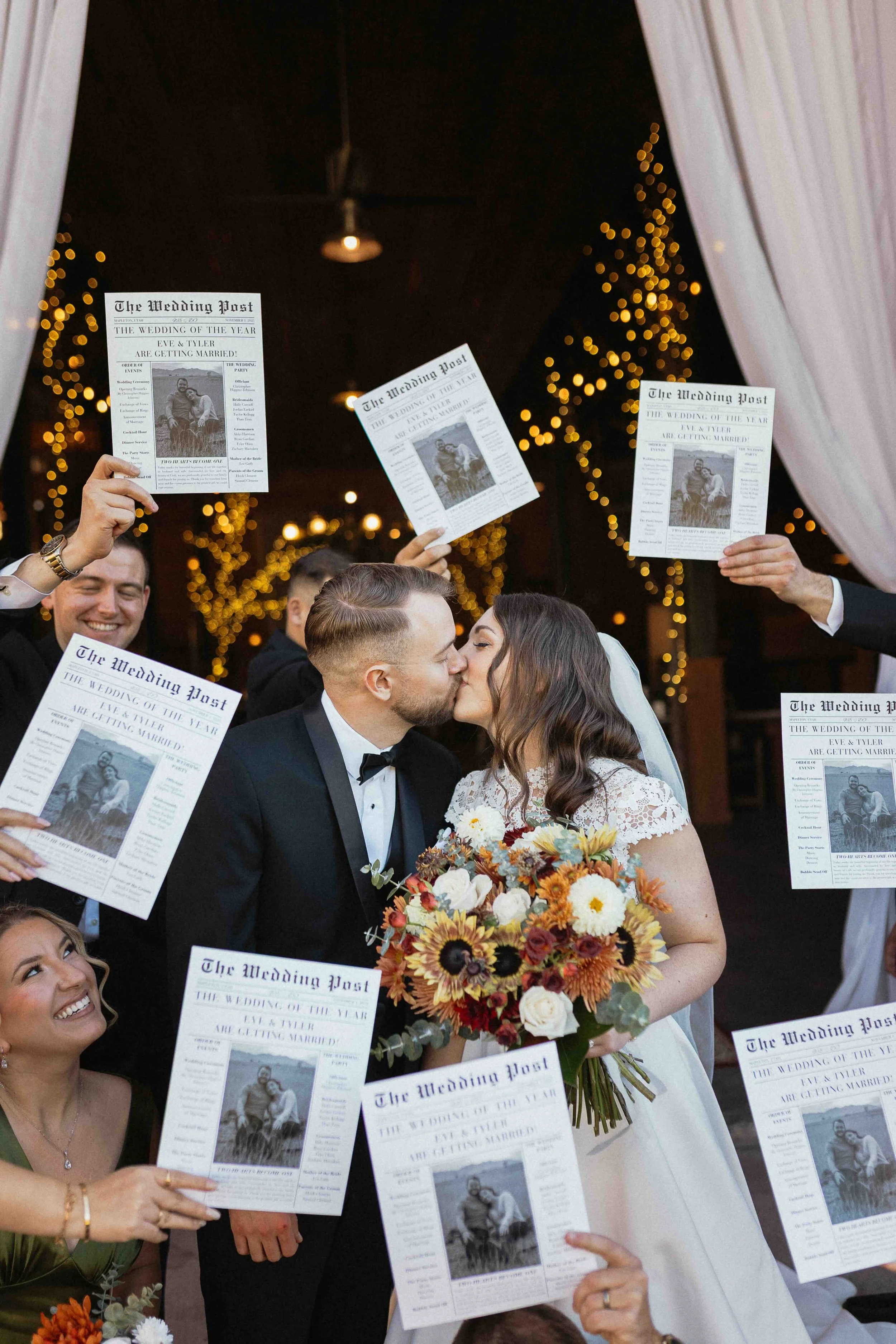 Bride and groom kissing surrounded by their bridal party holding newspapers celebrating the couple!