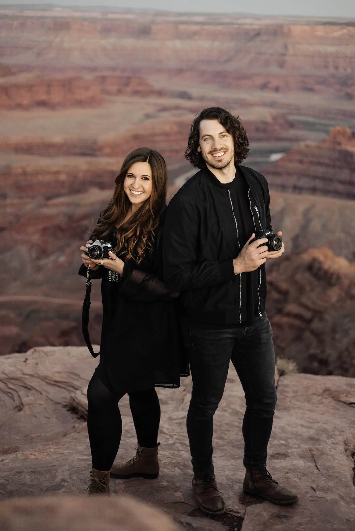 A husband and wife, photo and video team smiling and dressed in black while holding their cameras in front of red rock.