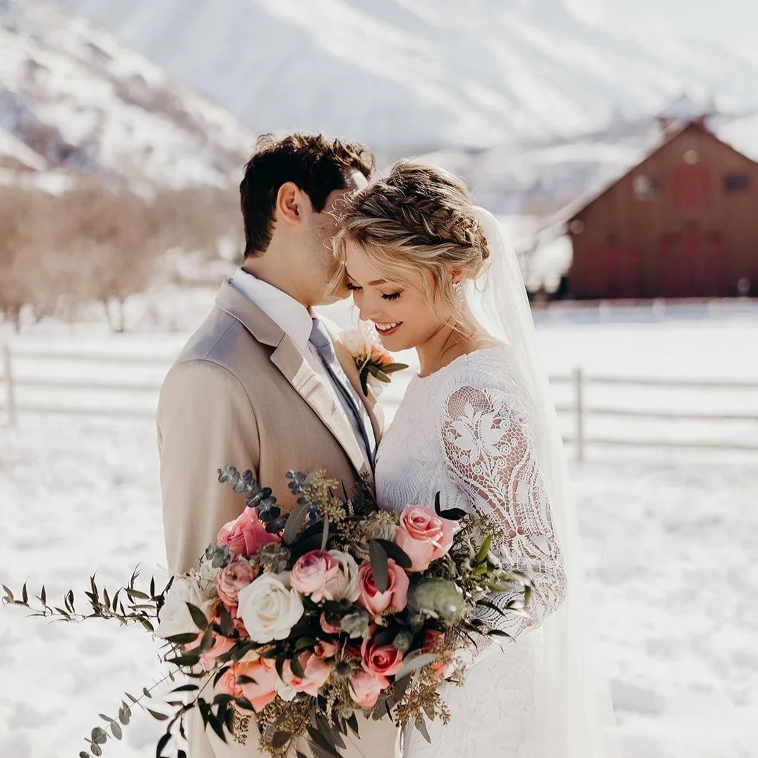 Bride and groom standing close in a snowy outdoor setting, with the bride holding a large bouquet of pink and white roses. The bride is wearing a white lace wedding dress, and the groom is in a light-colored suit. Both are smiling and leaning their foreheads together.