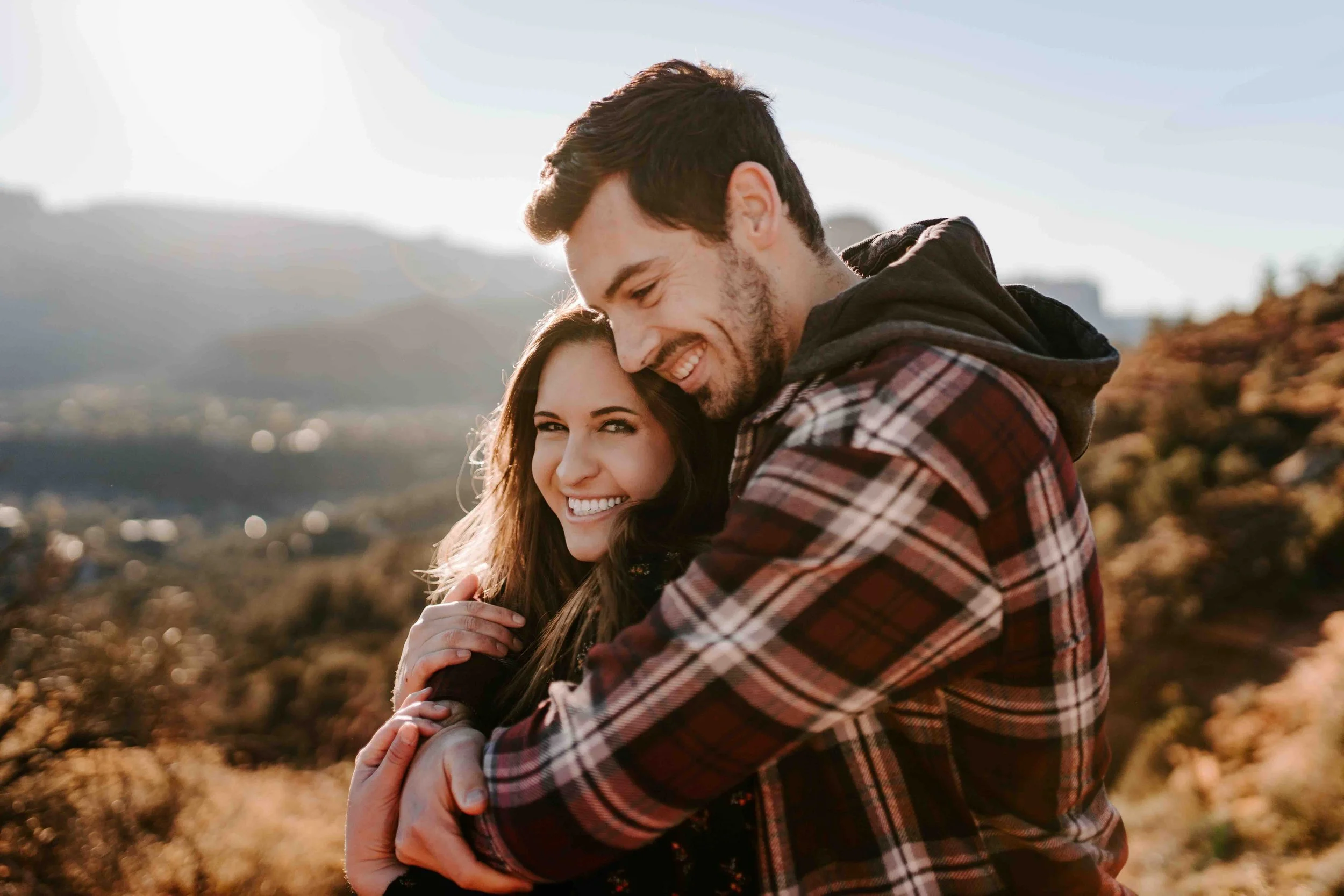 A happy couple hugging outdoors in a scenic natural setting, with mountains and a partly cloudy sky in the background.