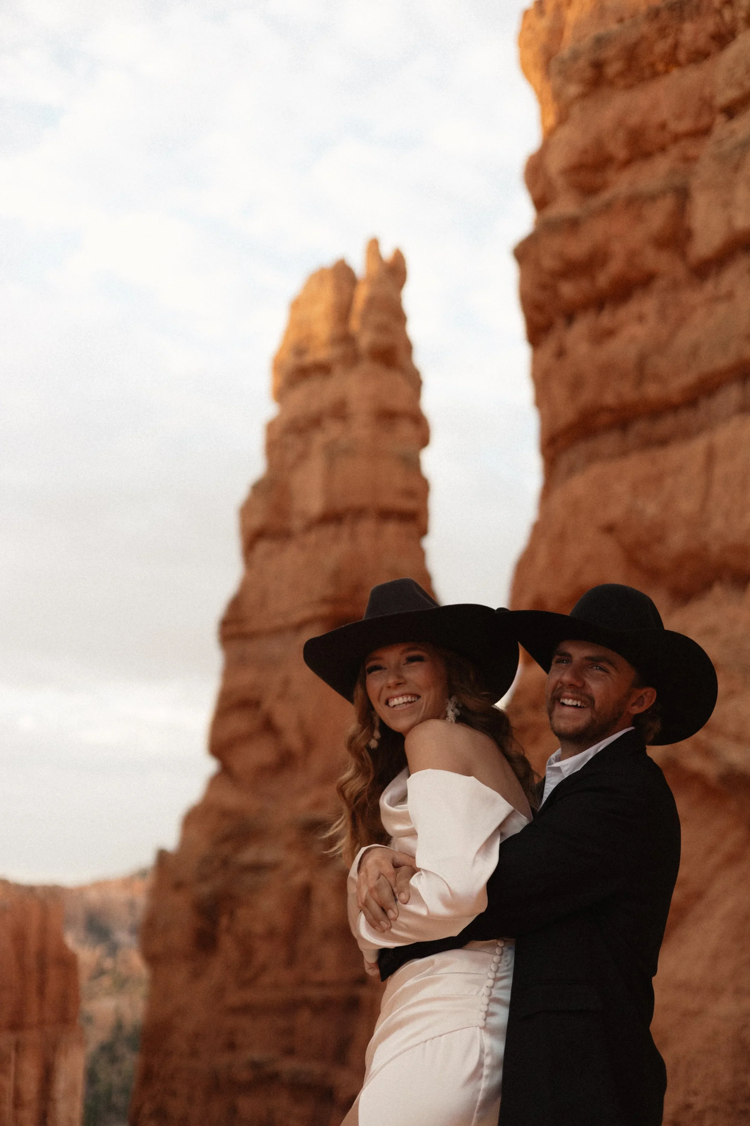 A smiling couple dressed in formal attire, with hats, stands in front of red rock formations in a desert landscape.
