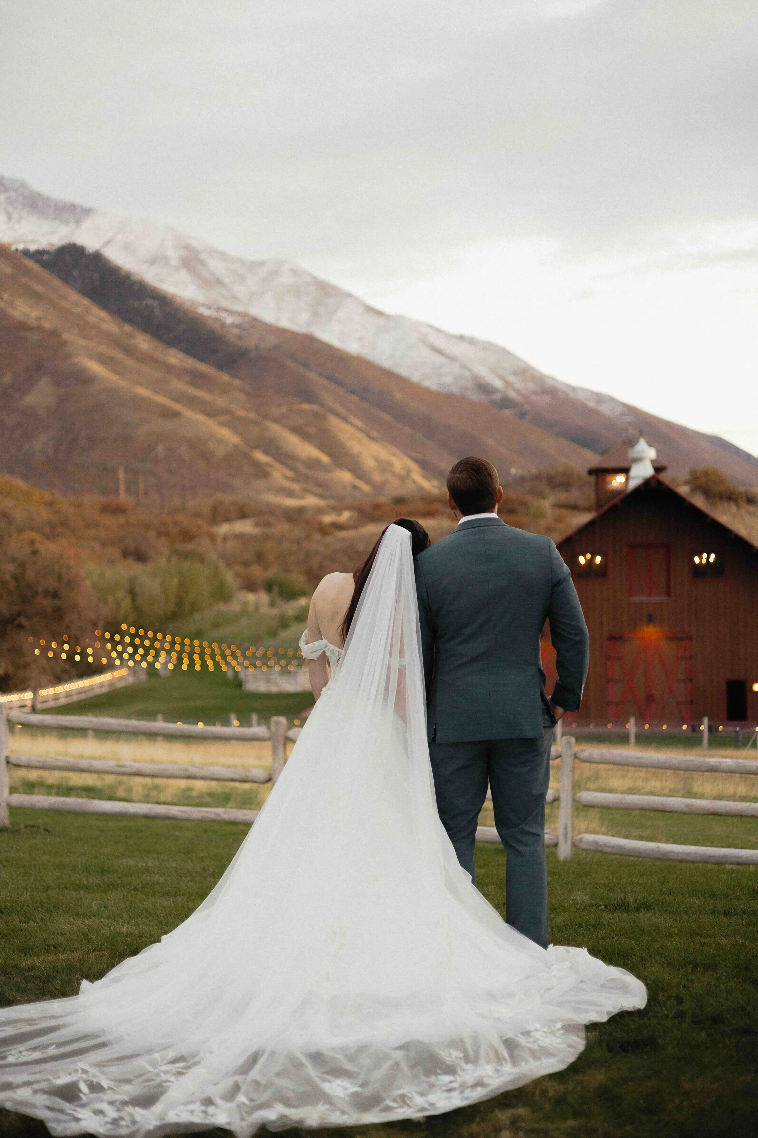 Bride and groom leaning on one another and taking in the view of the mountains and a lit up barn.